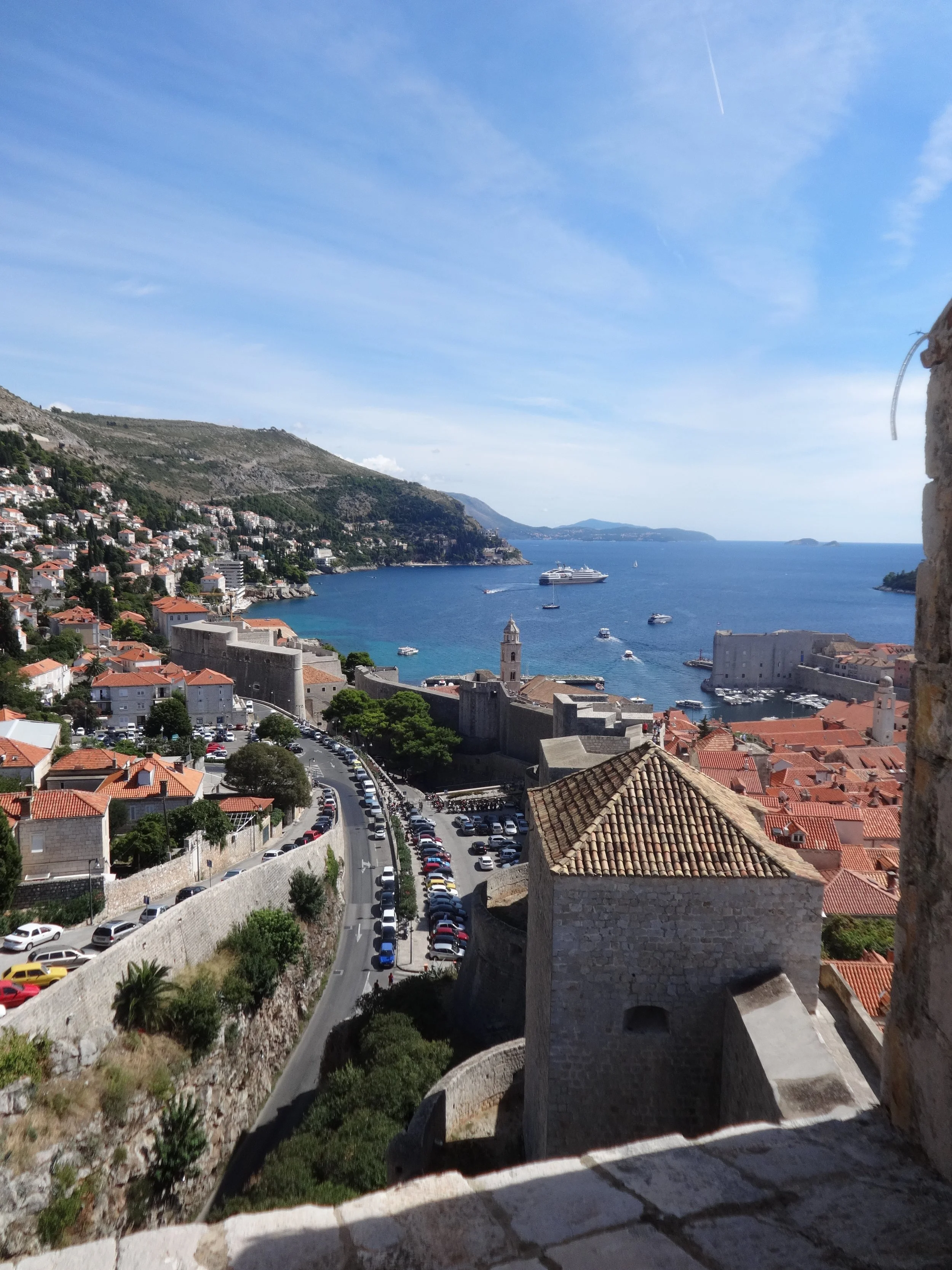 A coastal city with red-tiled roofs, a historic stone fortress, and a harbor filled with boats, overlooking the sea with a cruise ship in the water and green hills in the background.