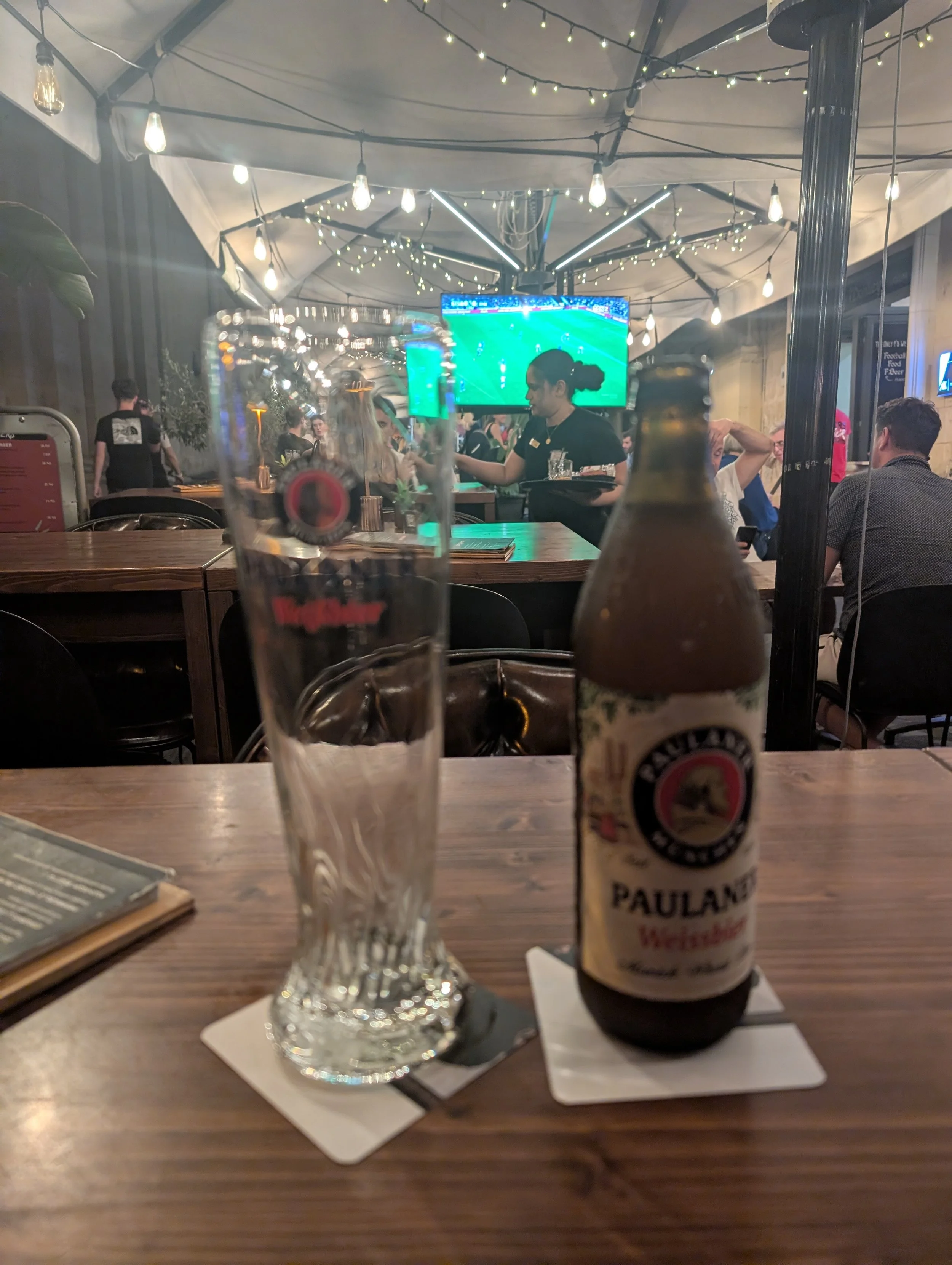A bar or restaurant setting with people dining, a large TV screen showing a soccer game, and decorative string lights. In the foreground, there is a clear glass with a logo, a bottle of beer labeled "Paulaner," and a menu on a wooden table.