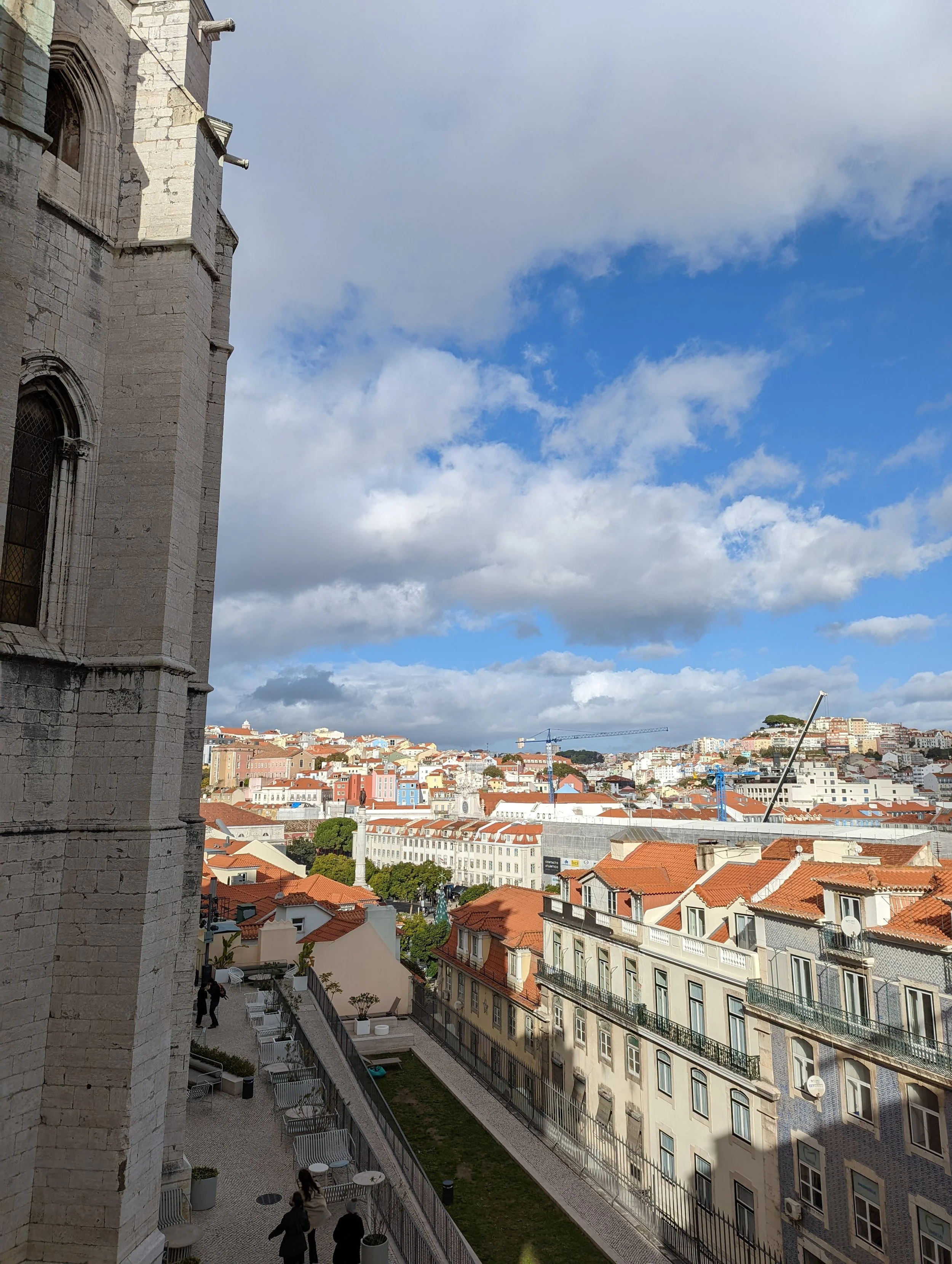 View of a cityscape under a partly cloudy sky, with a historic stone building on the left side and a mix of residential and commercial buildings with red and orange rooftops, some under construction cranes, and a small garden area at the bottom.