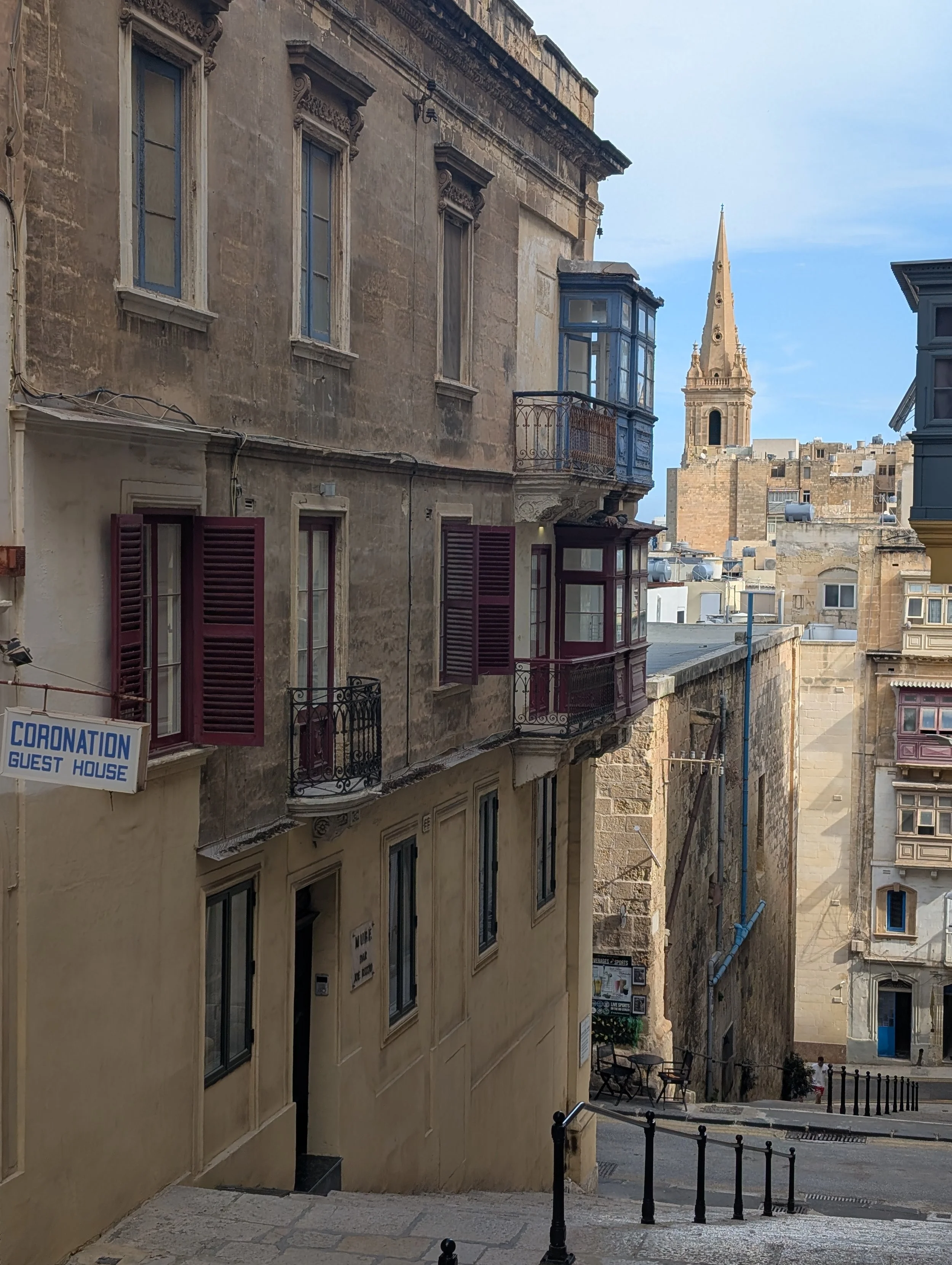 Narrow street with stone buildings, some with colorful balconies, a tower with a spire in the background, and a blue sky.