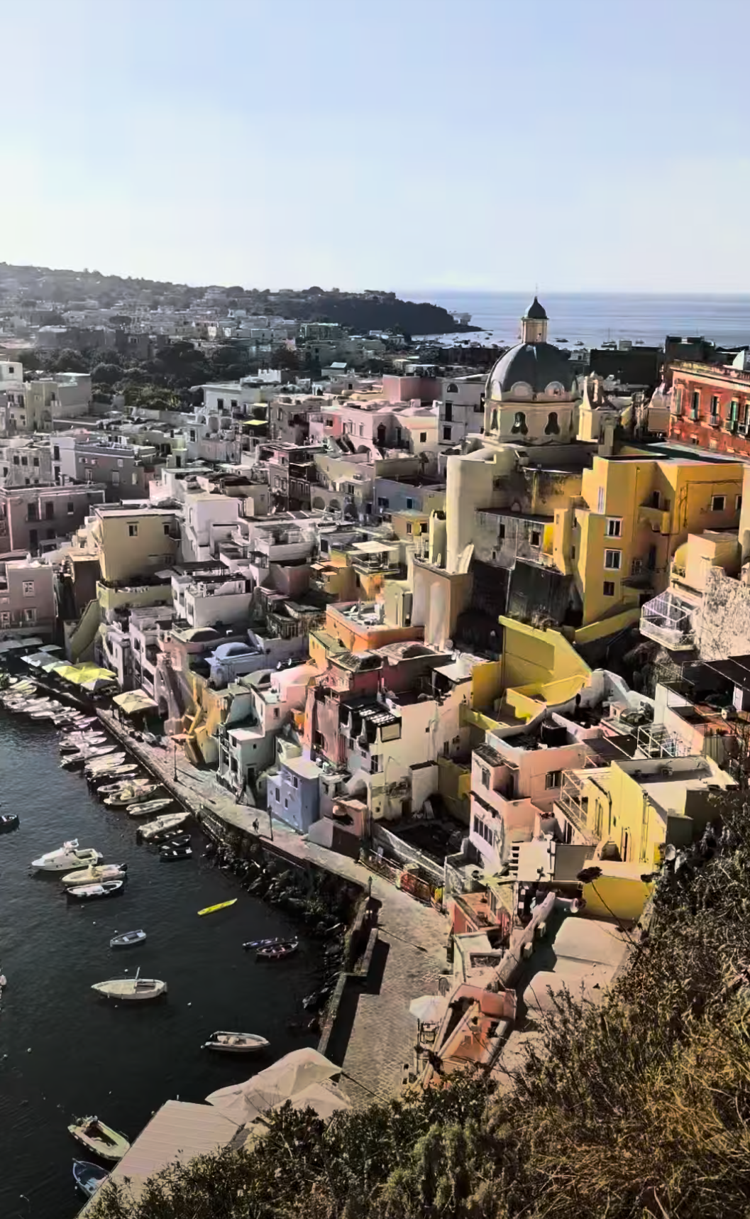 Colorful buildings along the waterfront with boats docked in the water, overlooking a scenic coastline with hills and the sea in the background.
