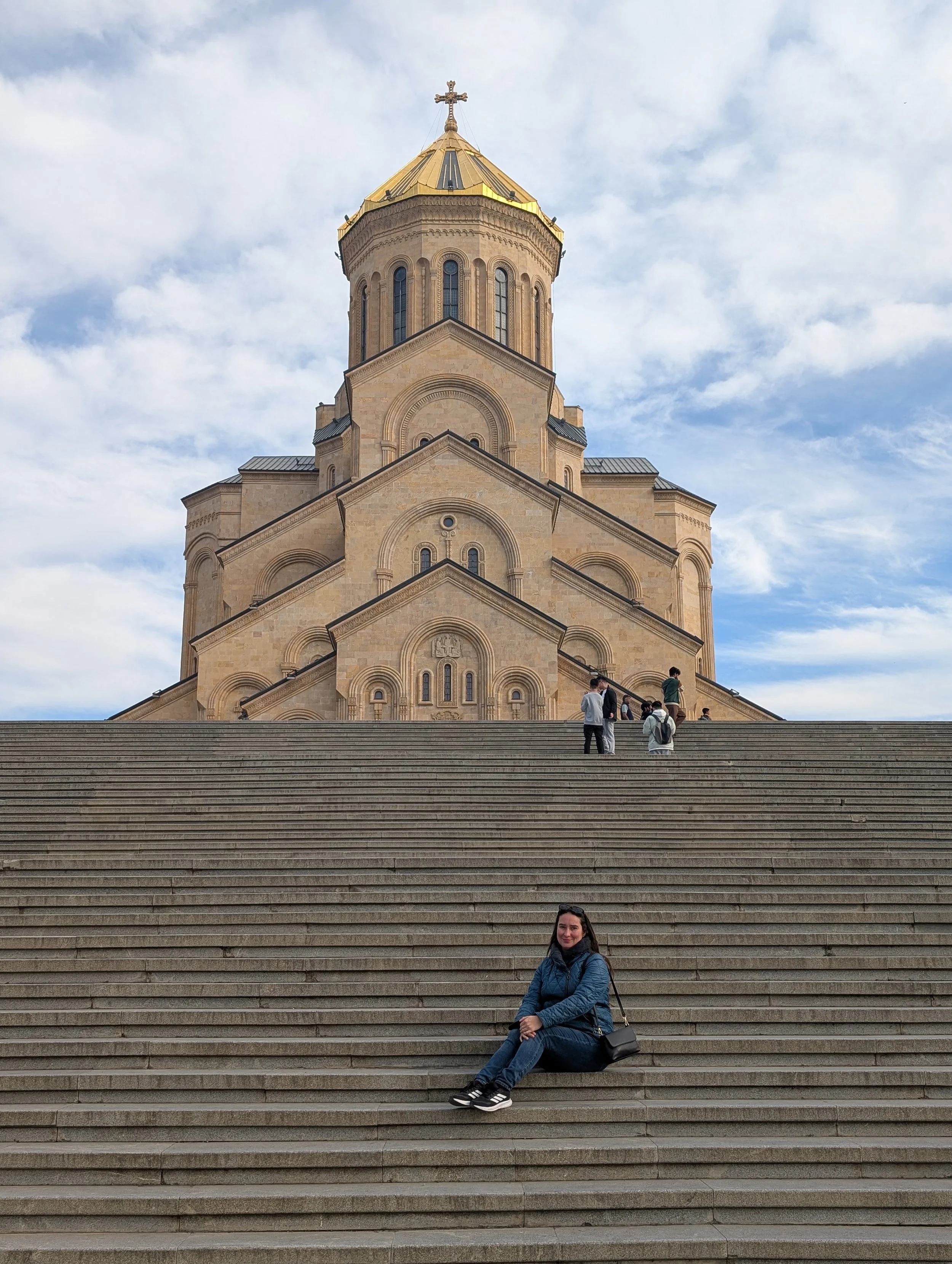 A woman sitting on wide staircase steps in front of a church with a tall, ornate tower topped with a golden dome and a cross. Several people stand or walk on the steps, and the sky is partly cloudy.