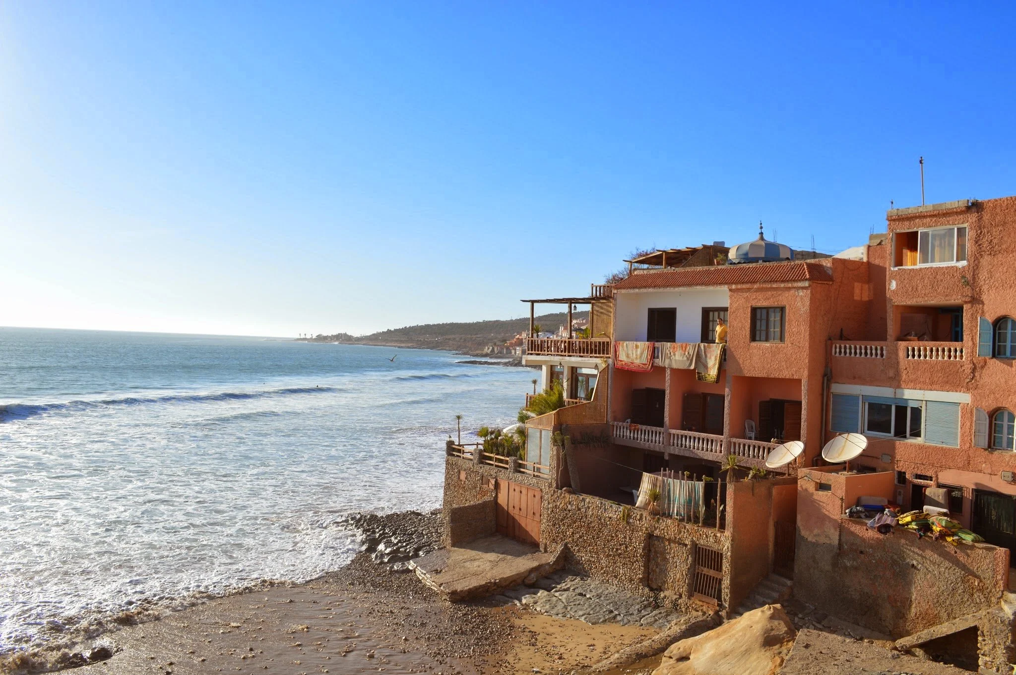 Beachside buildings with balconies and laundry hanging, overlooking the ocean with waves and a clear blue sky.