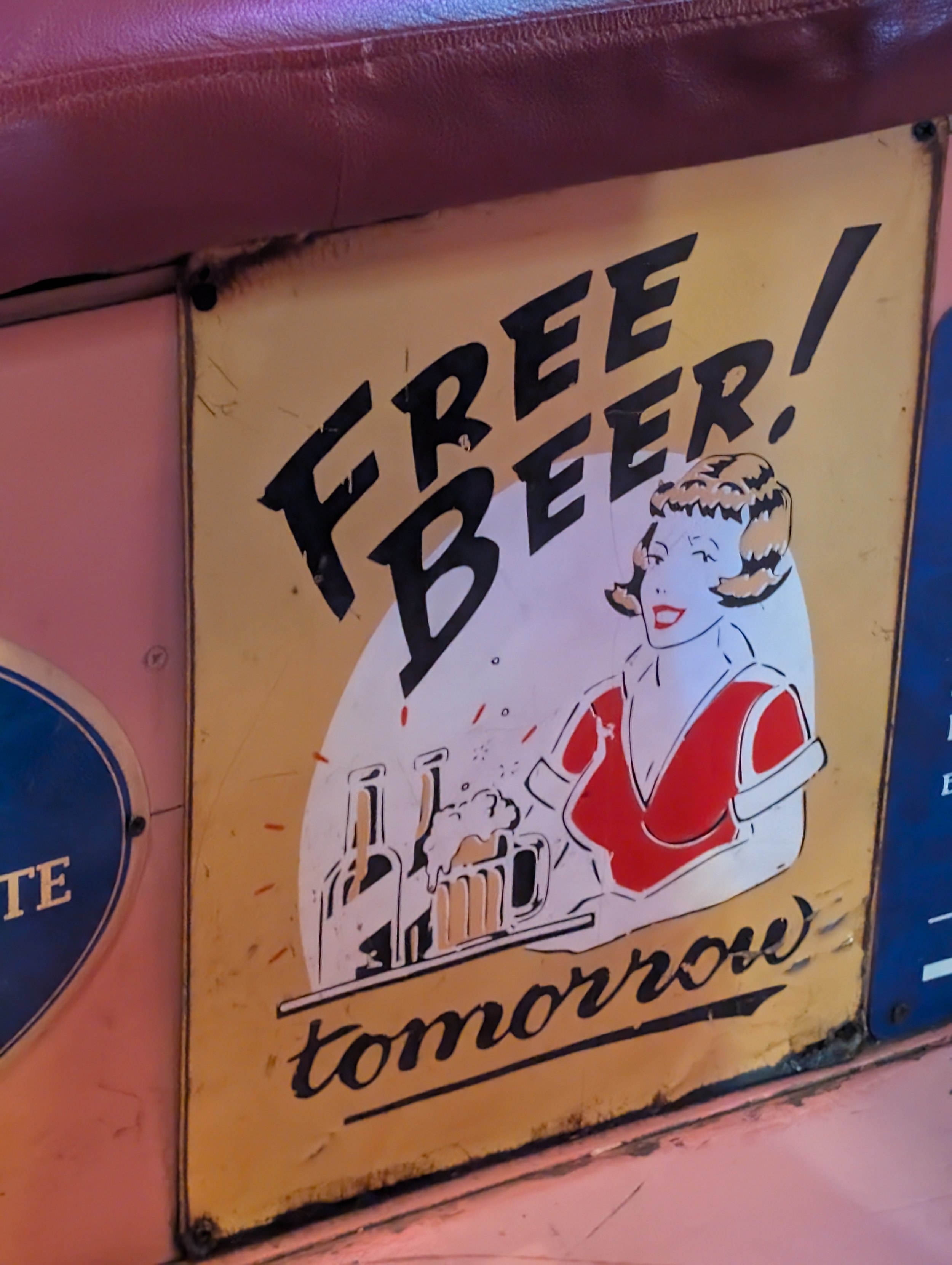 Vintage sign featuring a cartoon woman with blonde hair and red lipstick, wearing a red and white sports jersey, with the words "FREE BEER!" in large black lettering above her.