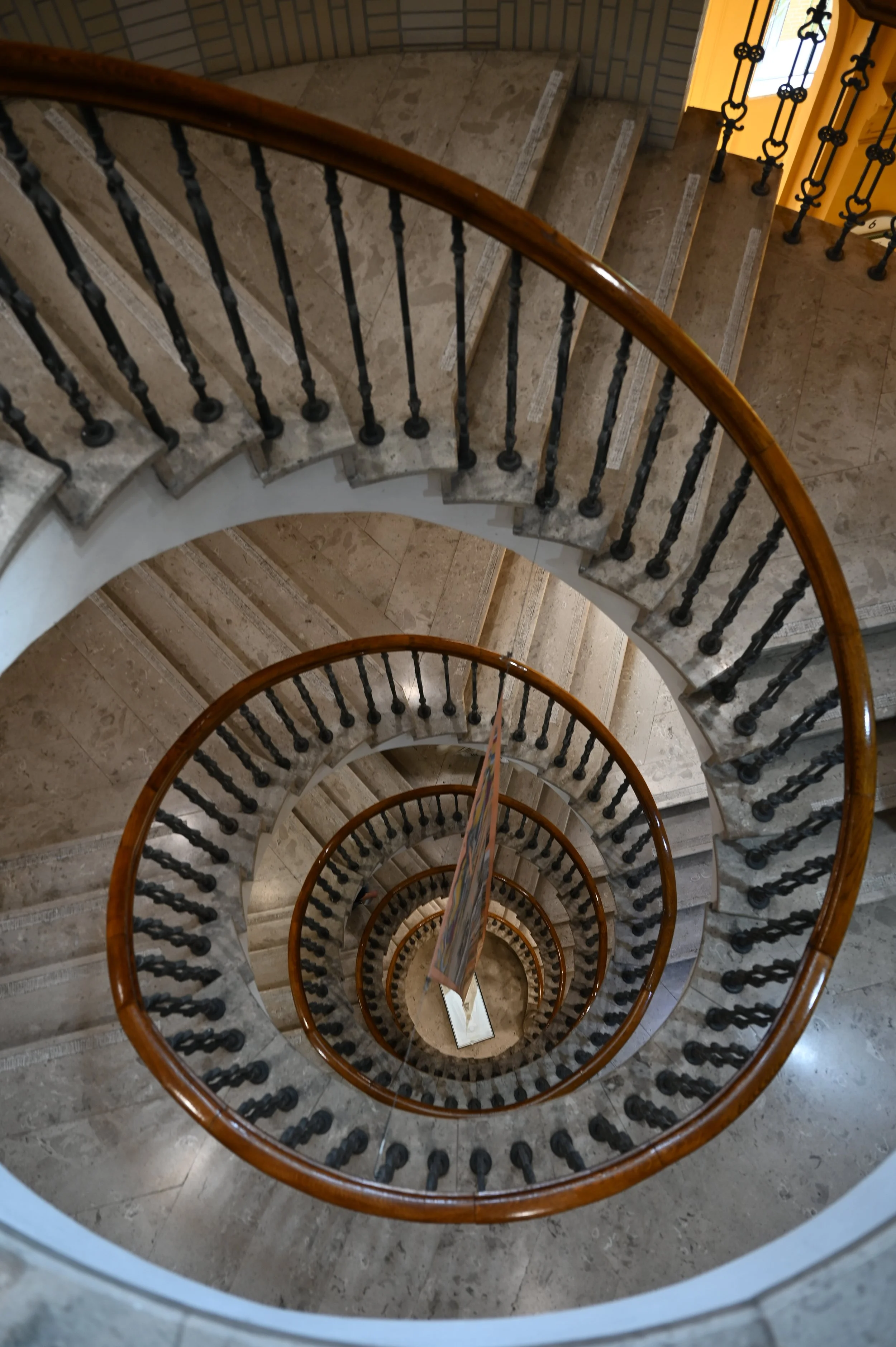 Spiral staircase viewed from above, with a wooden handrail and black metal balusters, descending through multiple floors with beige marble flooring.