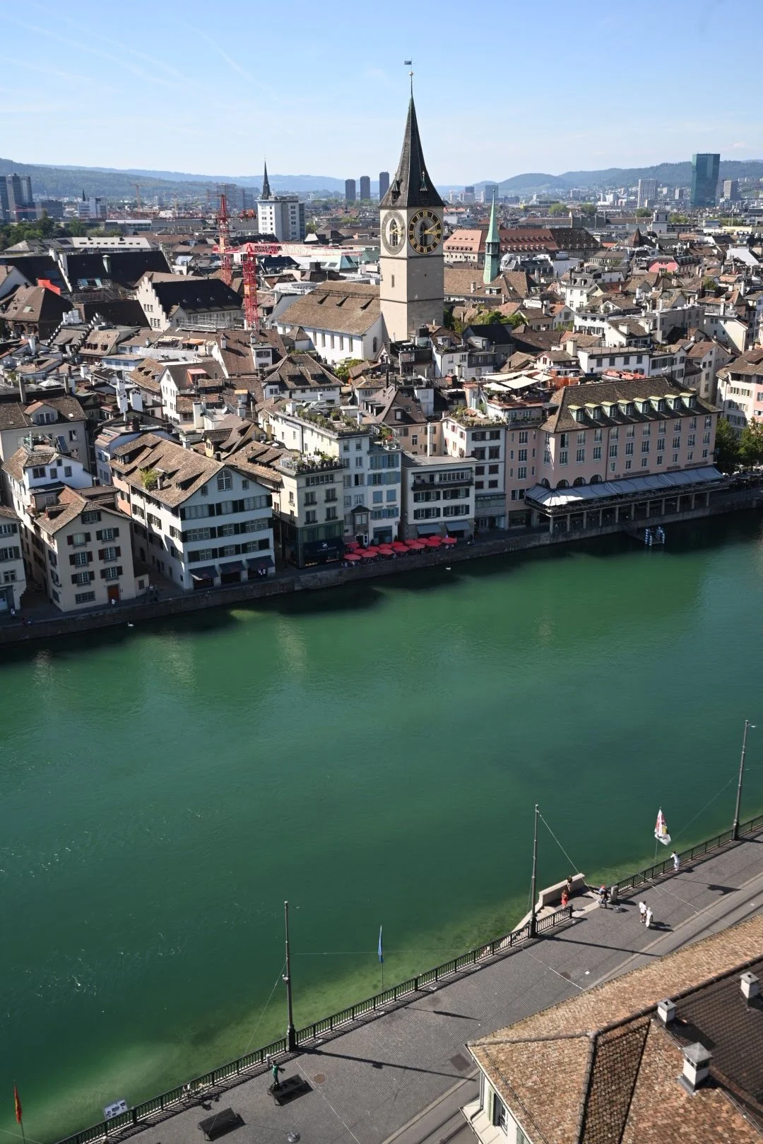 Aerial view of a city with a river in the foreground, historic buildings, a prominent church with a tall clock tower, and modern high-rise buildings in the background.