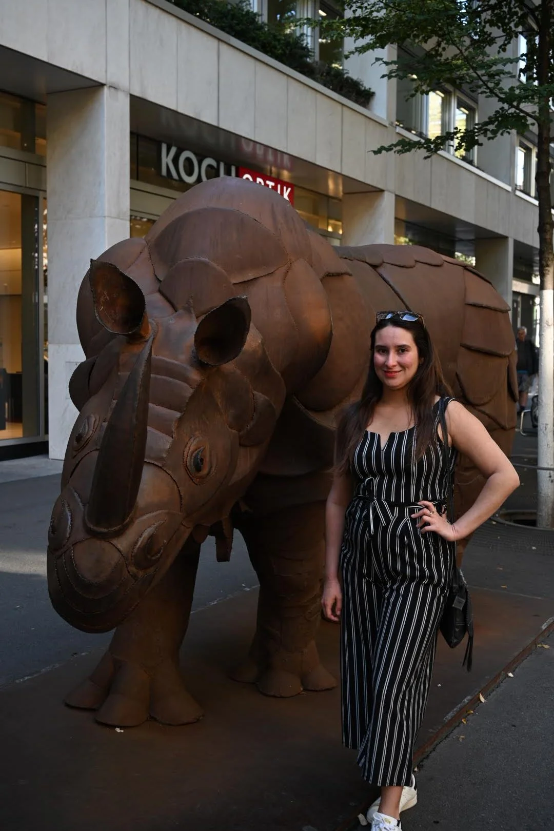 A woman with long brown hair, wearing a black and white striped jumpsuit, stands smiling next to a large, metal sculpture of a rhinoceros on a city street.