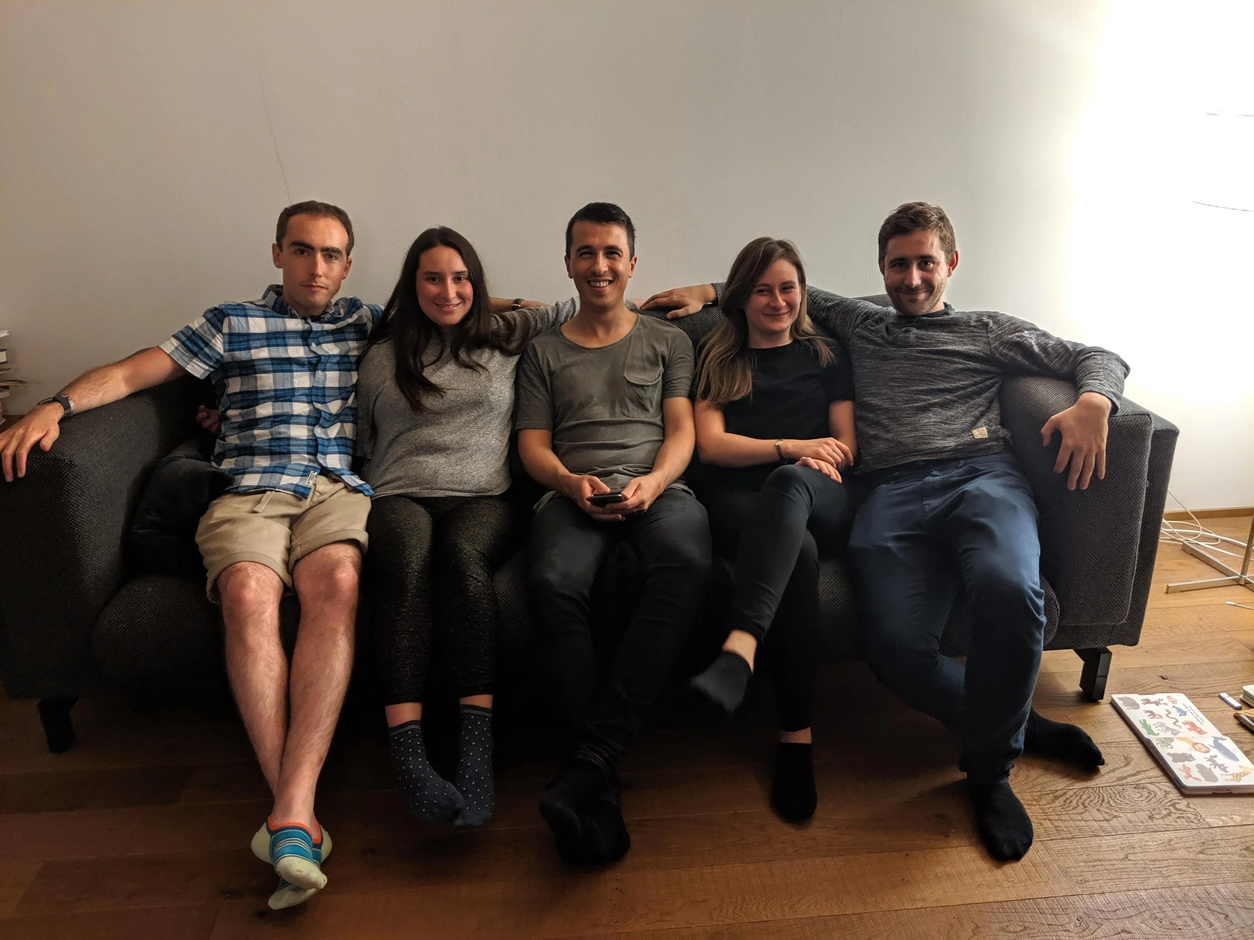 Six young adults sitting on a black couch in a living room, smiling and relaxing together. They are casually dressed, with some wearing shorts and others in T-shirts and sweatshirts. A board game is visible on the floor to the right.