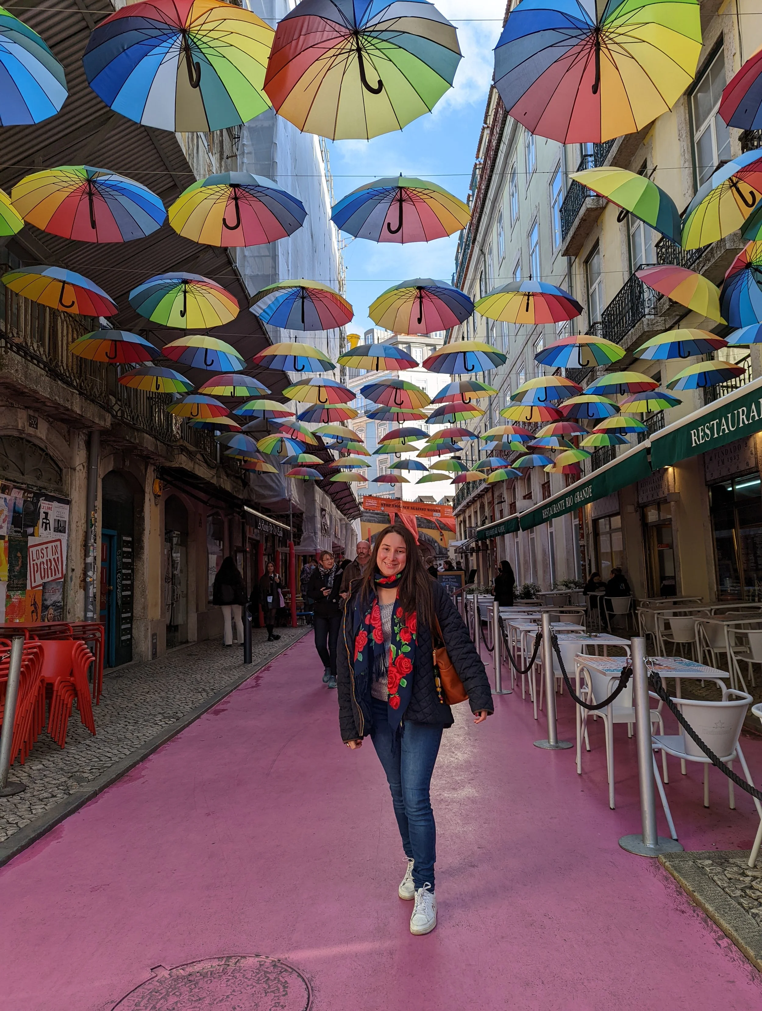 A woman standing on a pink pedestrian street with colorful umbrellas hanging overhead, smiling at the camera, with buildings lining the street and outdoor restaurant seating visible.