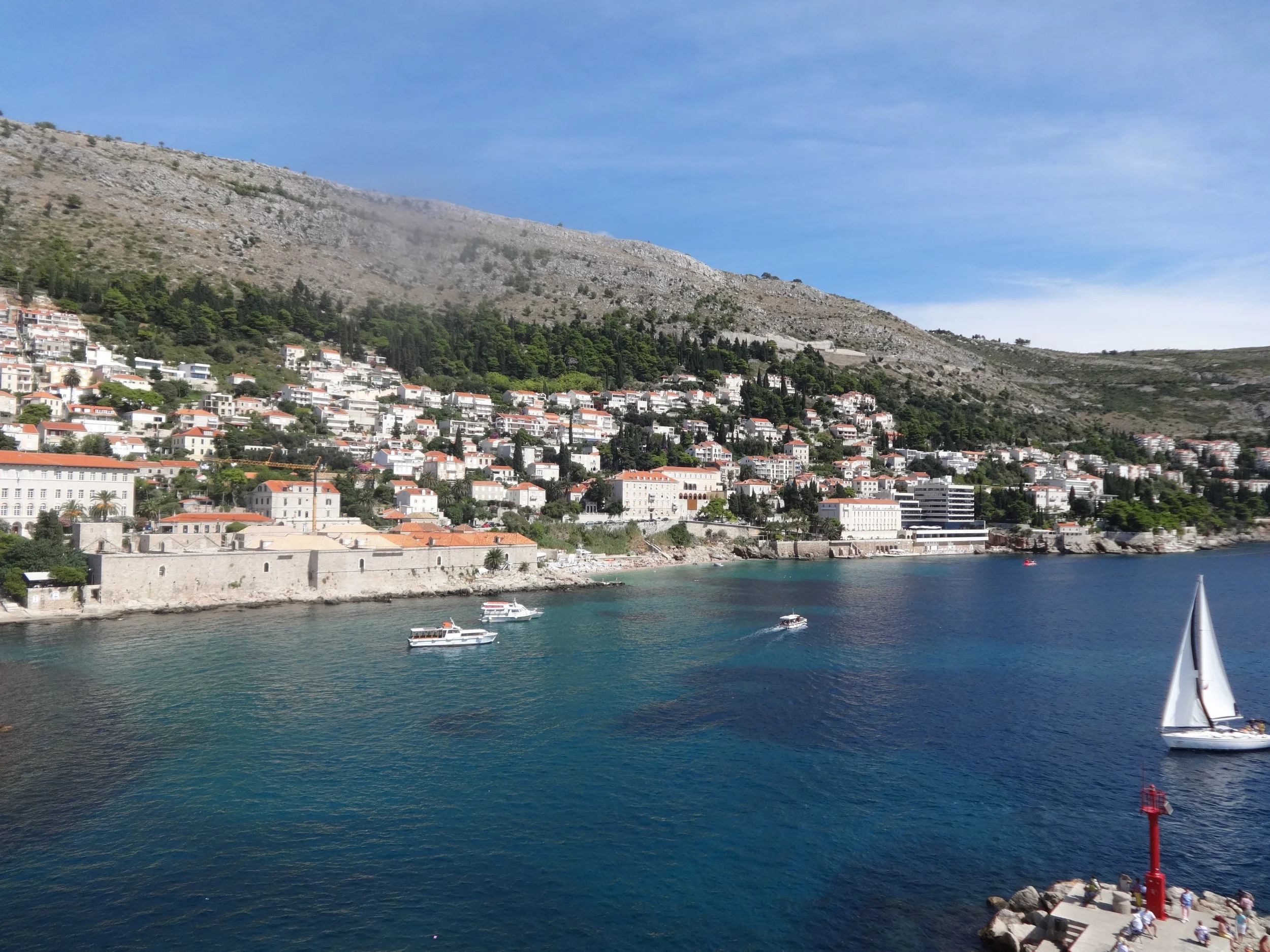 A coastal town with white buildings and terracotta roofs situated on the hillside, overlooking a body of water with boats, by a rocky shoreline under a clear blue sky.
