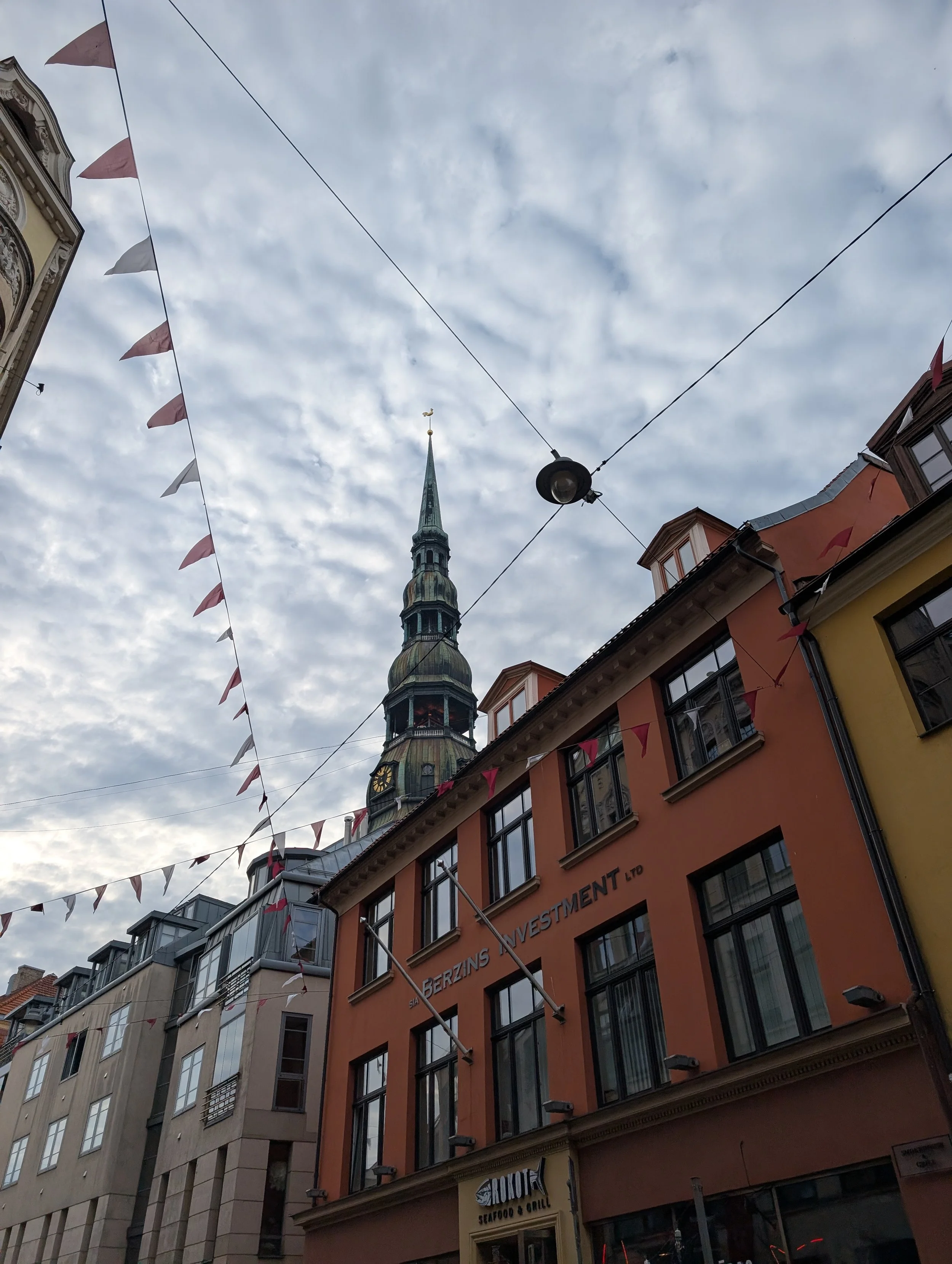 View of a street with colorful buildings, string flags, and a church spire against cloudy sky.