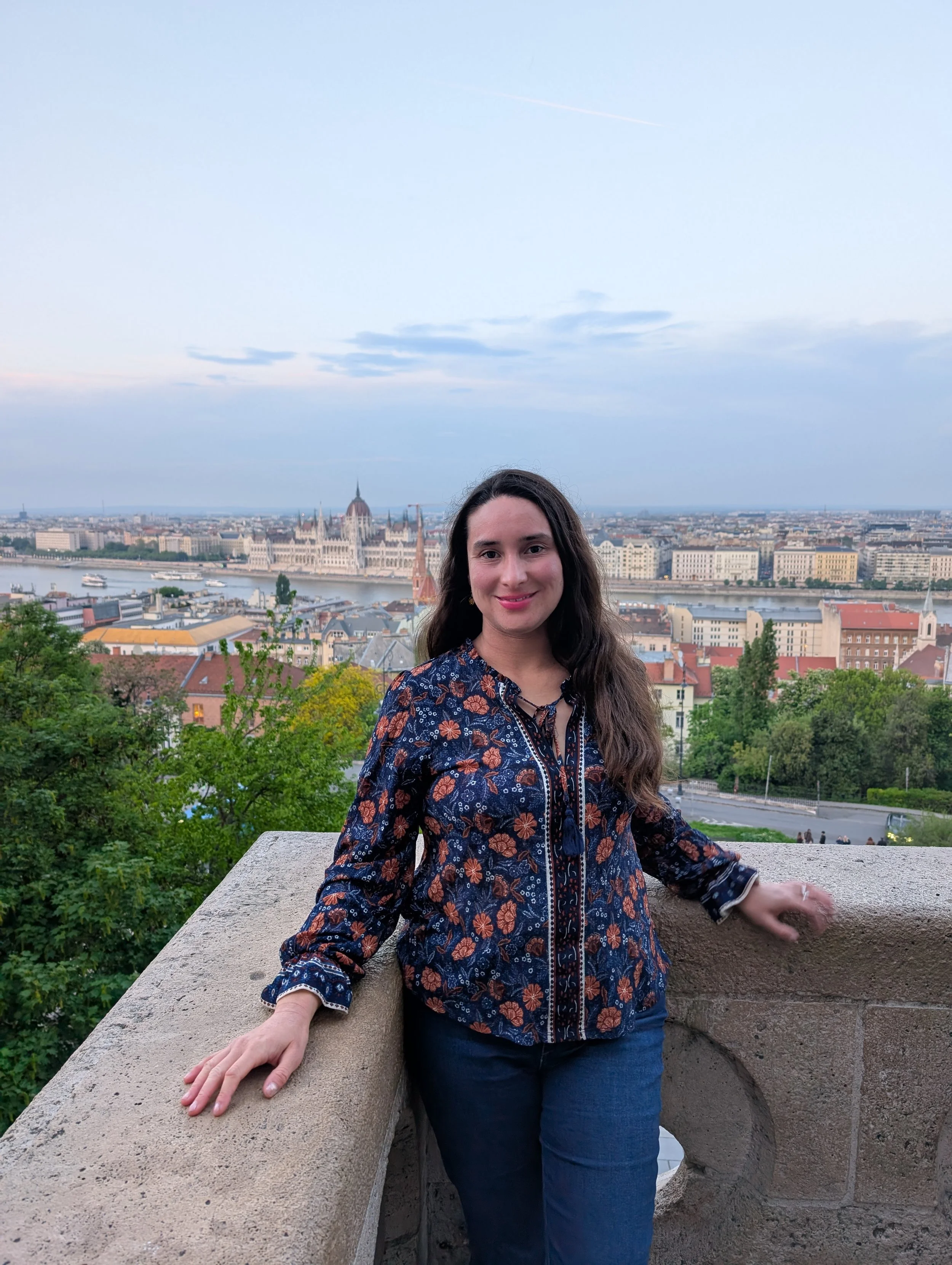 A woman with long dark hair and a floral-patterned jacket standing on a stone balcony with a cityscape and a river in the background, under a partly cloudy sky.