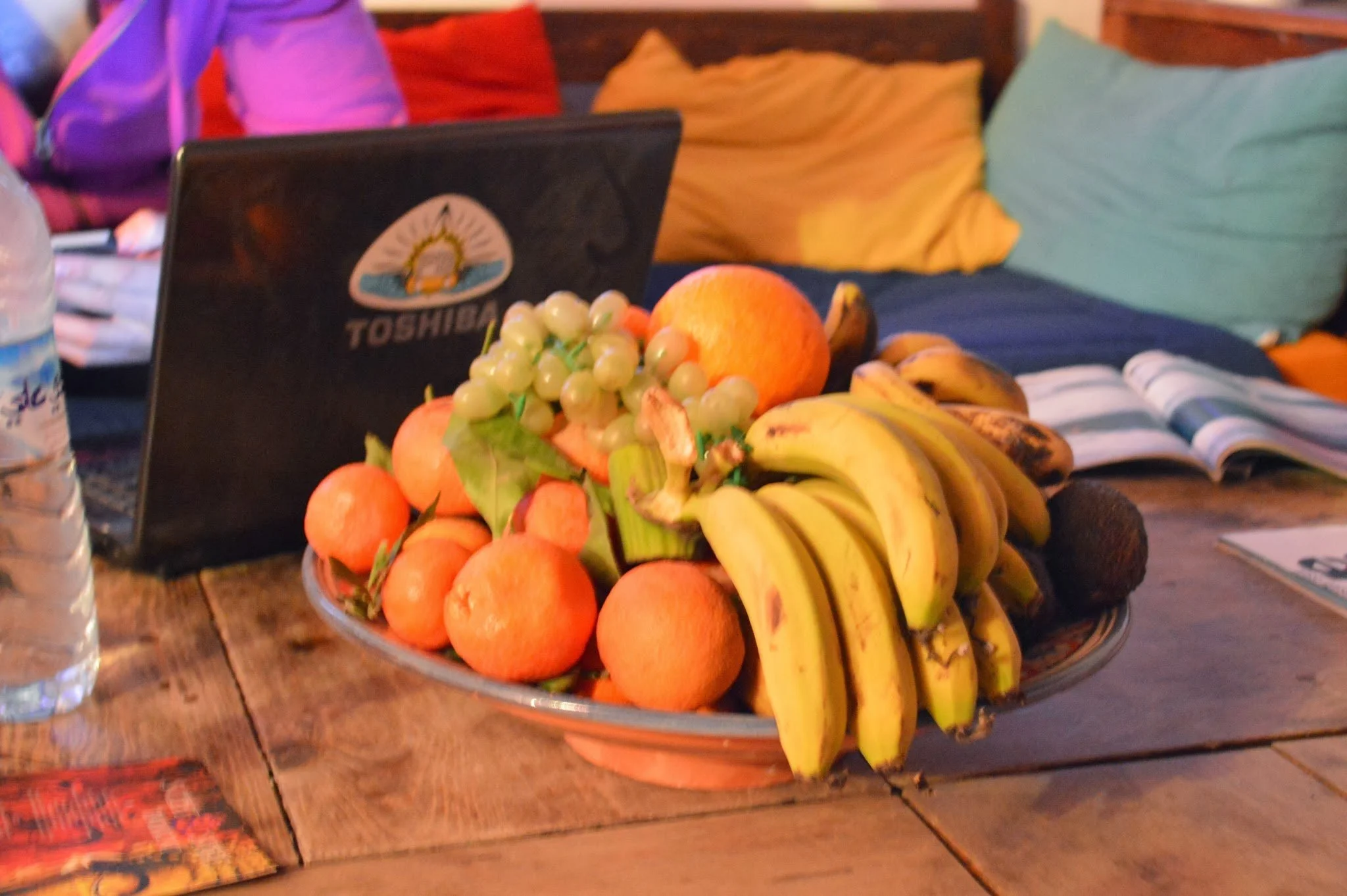 A glass bowl filled with oranges, green grapes, bananas, and pomegranate placed on a wooden table. In the background, there is a laptop with a sticker, bottles of water, and colorful pillows on a couch.
