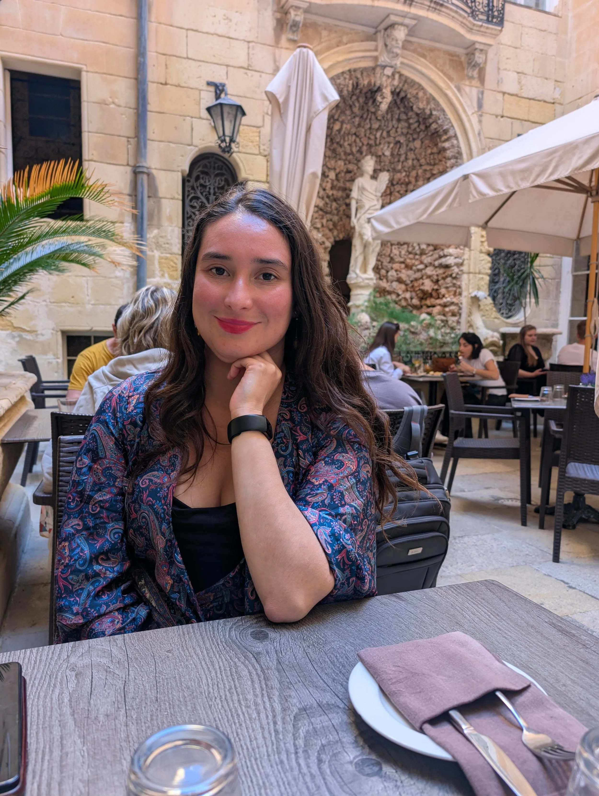 Woman with long dark hair and a colourful patterned top sitting at an outdoor restaurant table, smiling with her chin resting on her hand, surrounded by other diners and classic European architecture