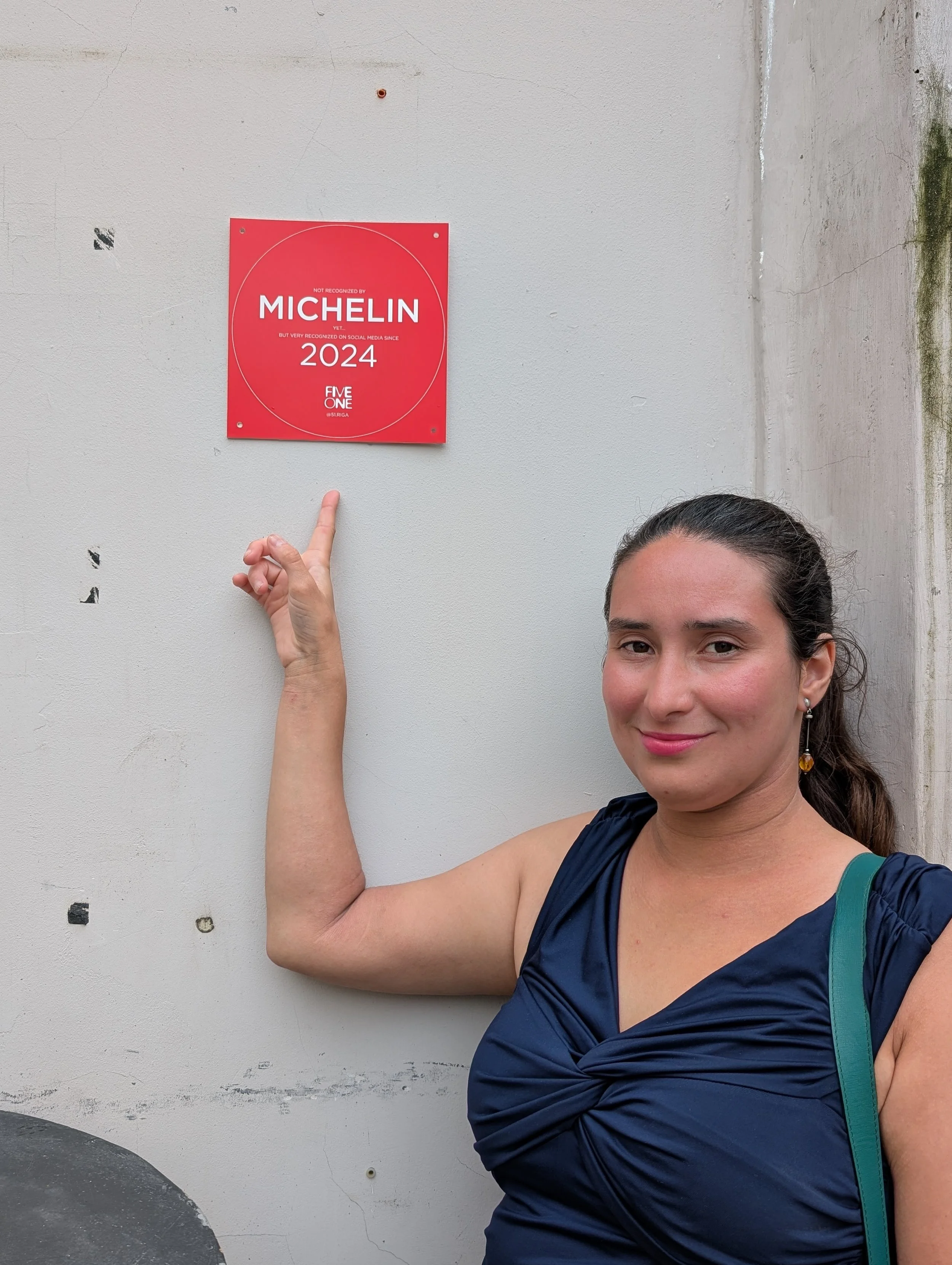 A woman with brown hair tied back, wearing a navy blue dress, earrings, and a turquoise shoulder bag, points at a red Michelin Guide sign on a white wall.
