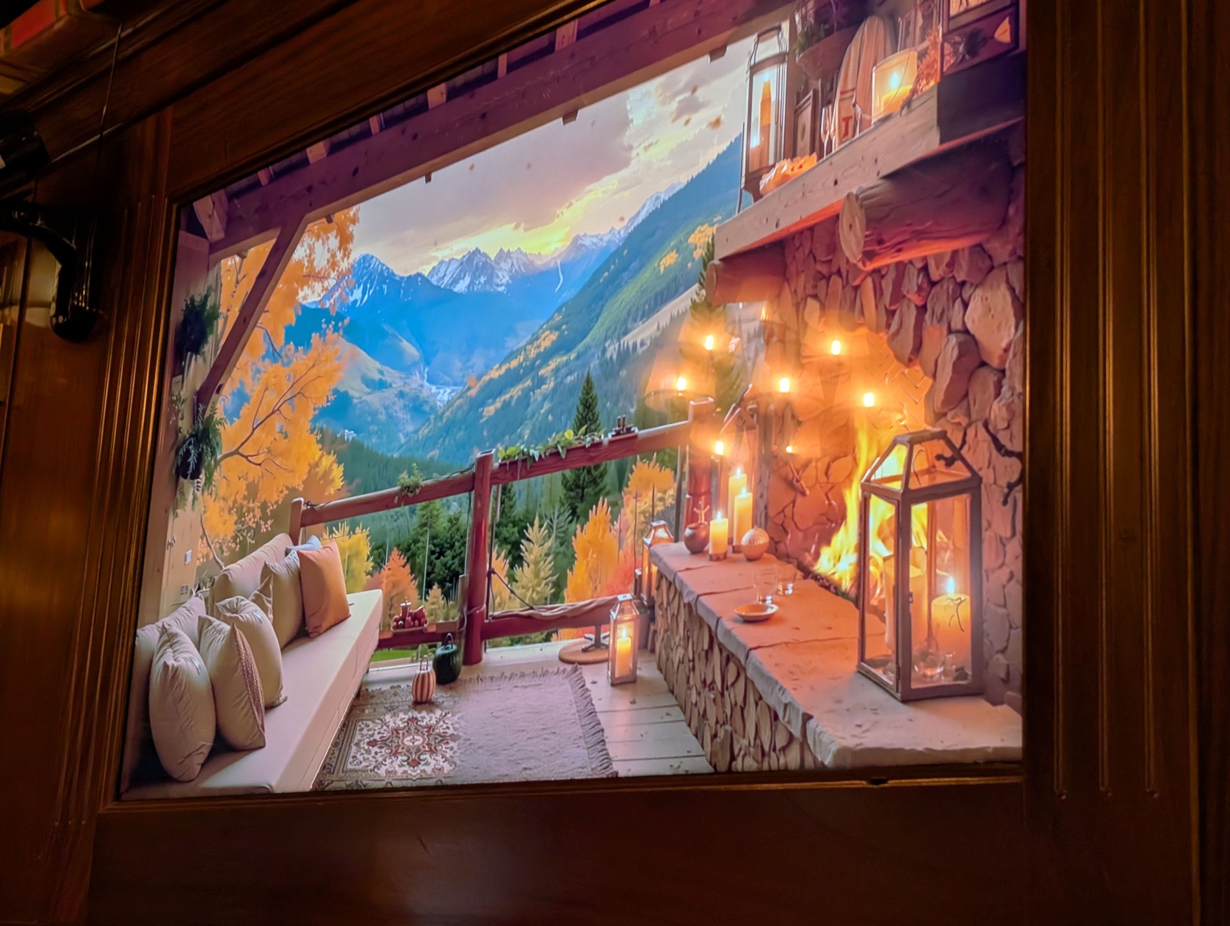 A cosy mountain cabin living room with a large window showcasing scenic mountains and trees with autumn foliage, decorated with candles, lanterns, and rustic decor.