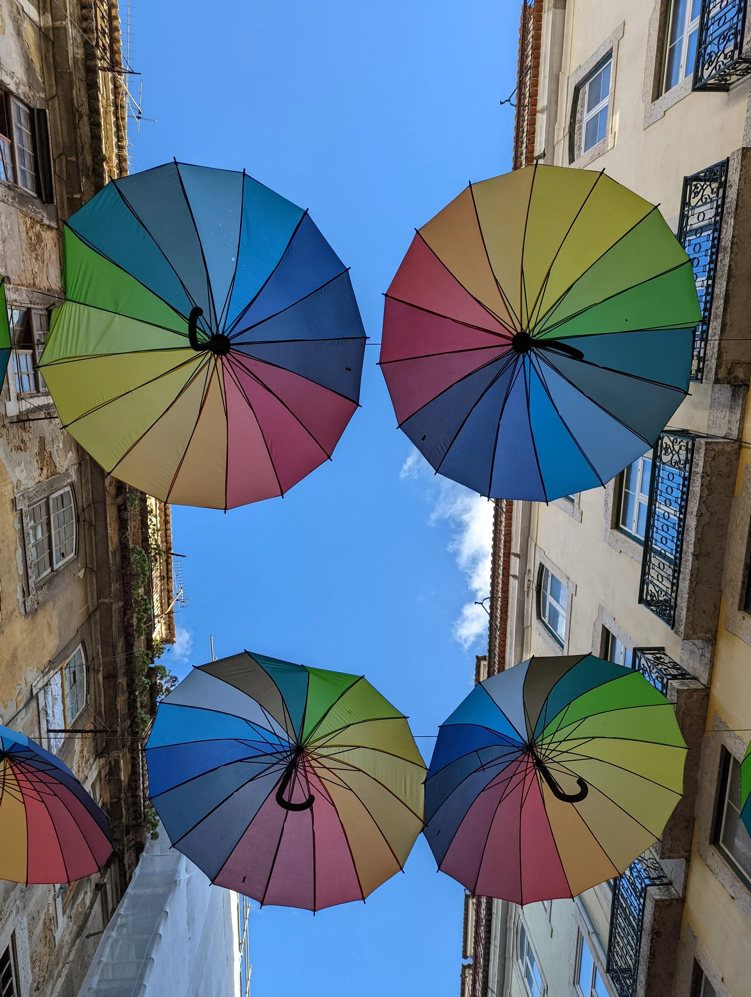 Colorful umbrellas suspended in the air against a bright blue sky, floating between old European-style buildings with balconies and windows.