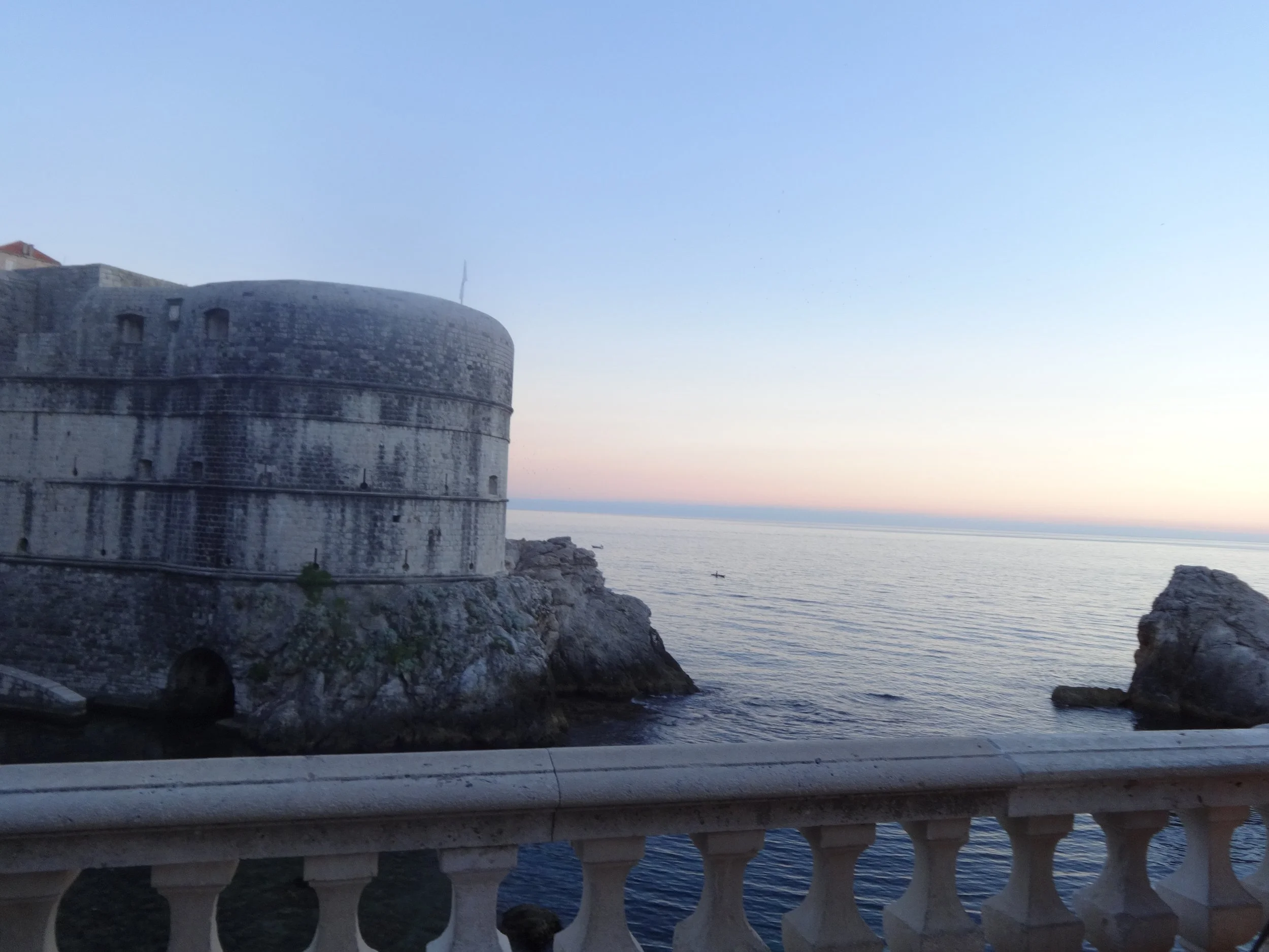 A historic stone fort or castle situated by the ocean, with a curved tower and small windows, viewed from a nearby road or walkway with a white balustrade railing in the foreground, during sunset or dusk.