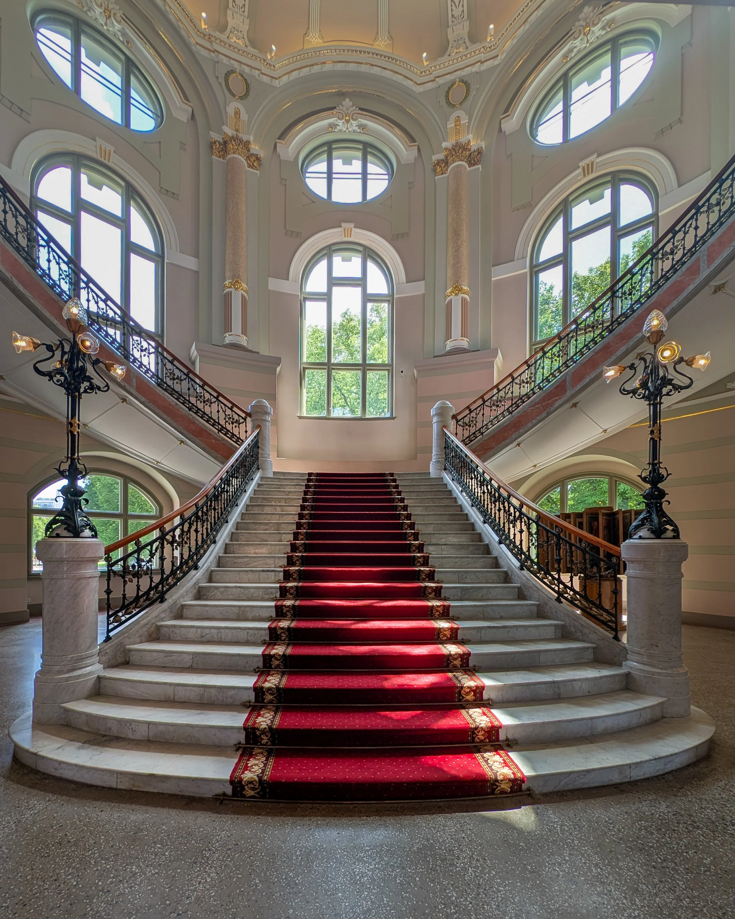 Grand staircase with red carpet runner, ornate black wrought iron railings, marble steps, and elegant chandeliers in a historic building with large arched windows and decorative columns.