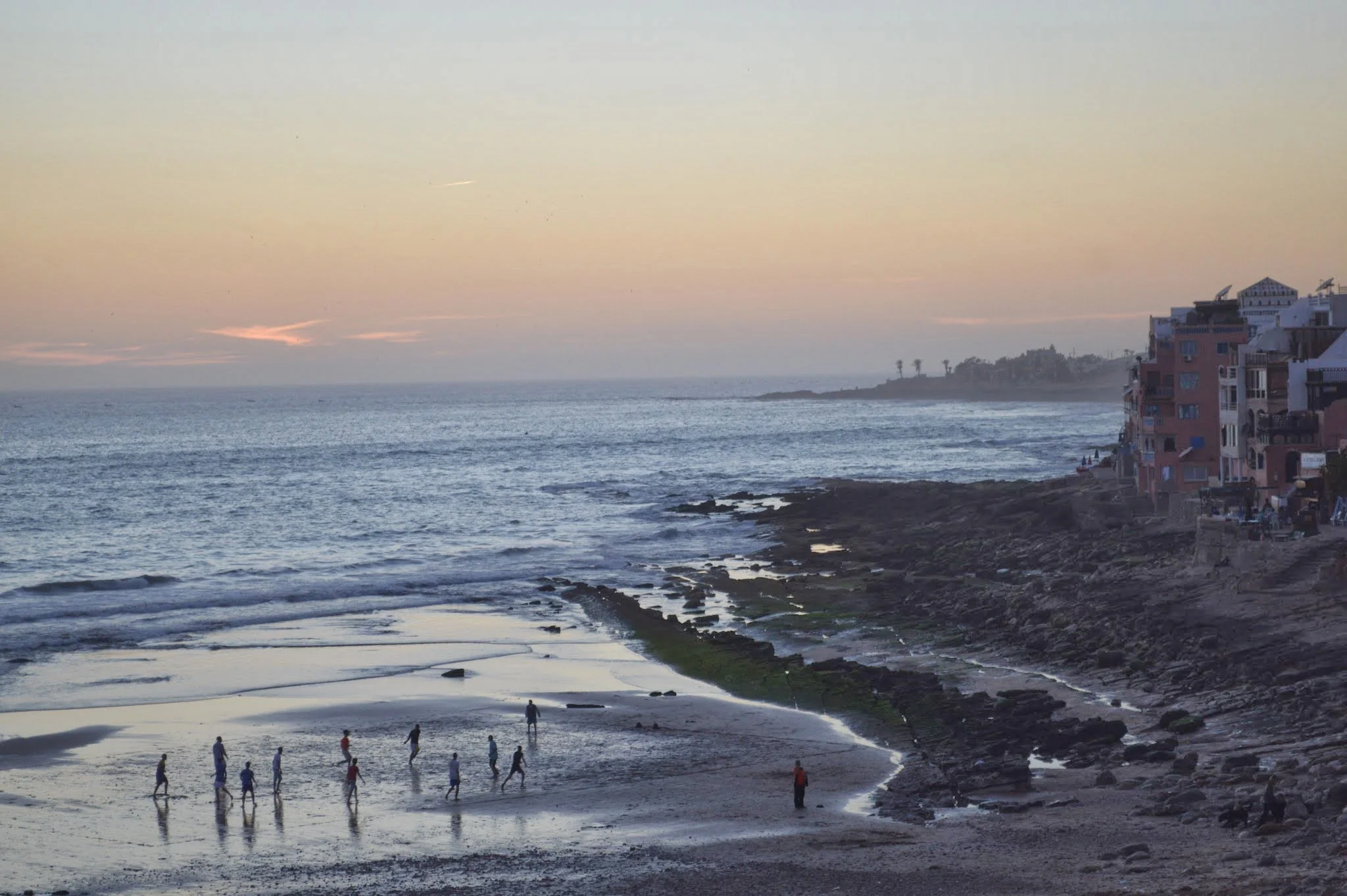 People walking along a beach at sunset with the ocean, rocky shoreline, and waterfront buildings in the background.