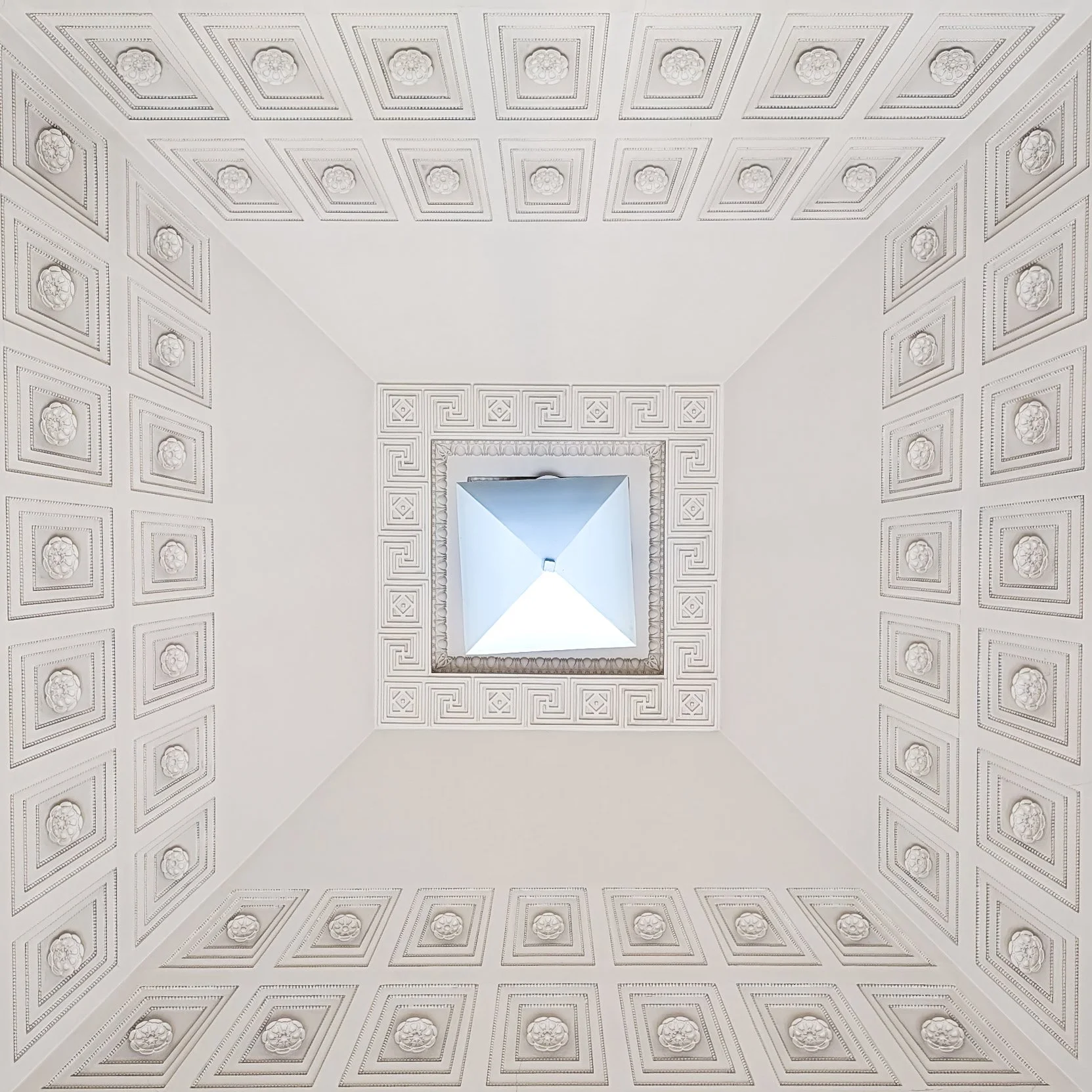 Looking up at a square ceiling with ornate floral and geometric patterns around a central skylight.