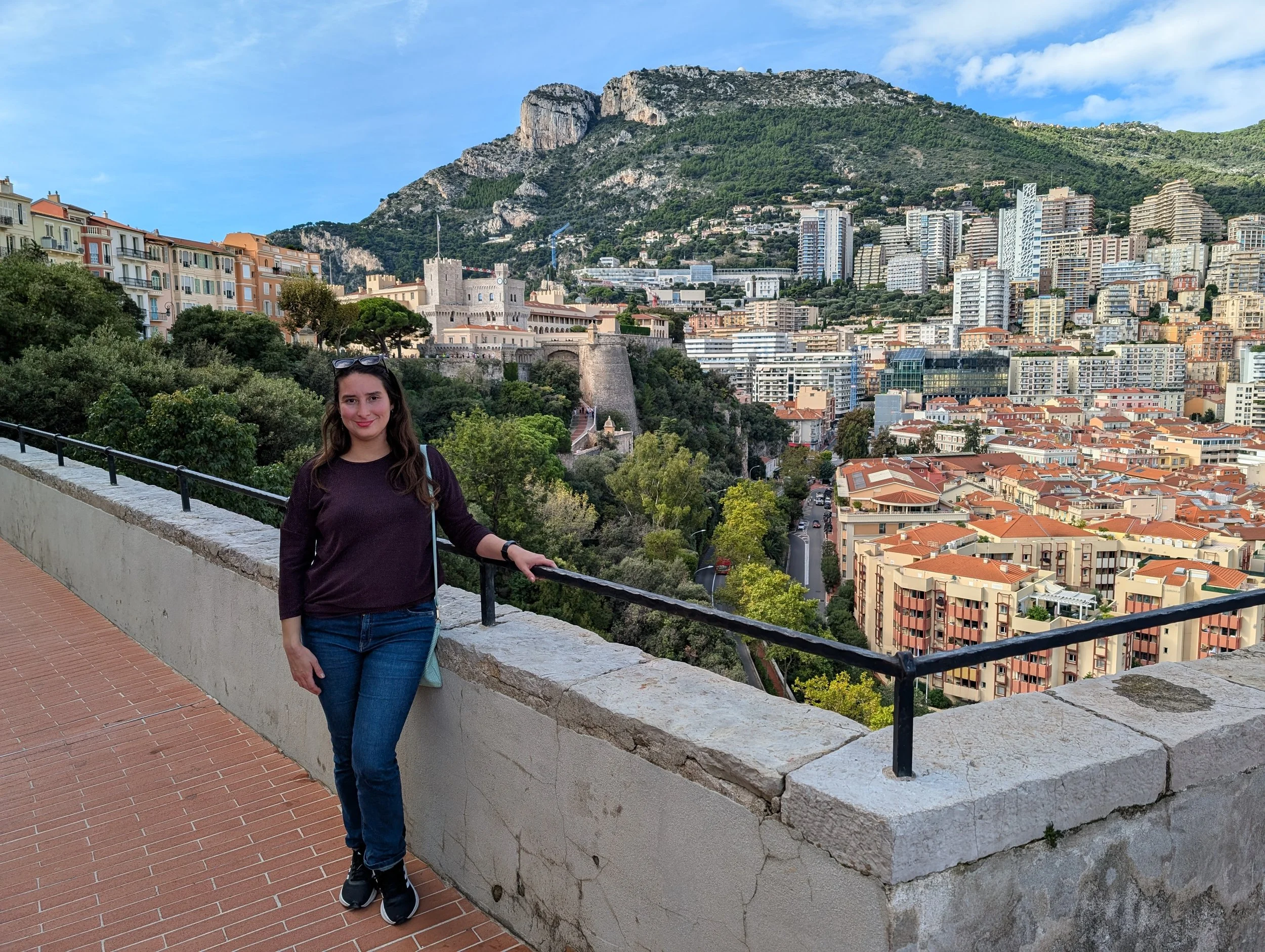 A woman standing near a stone wall overlooking a city with high-rise buildings and lush green mountains in the background on a clear day.