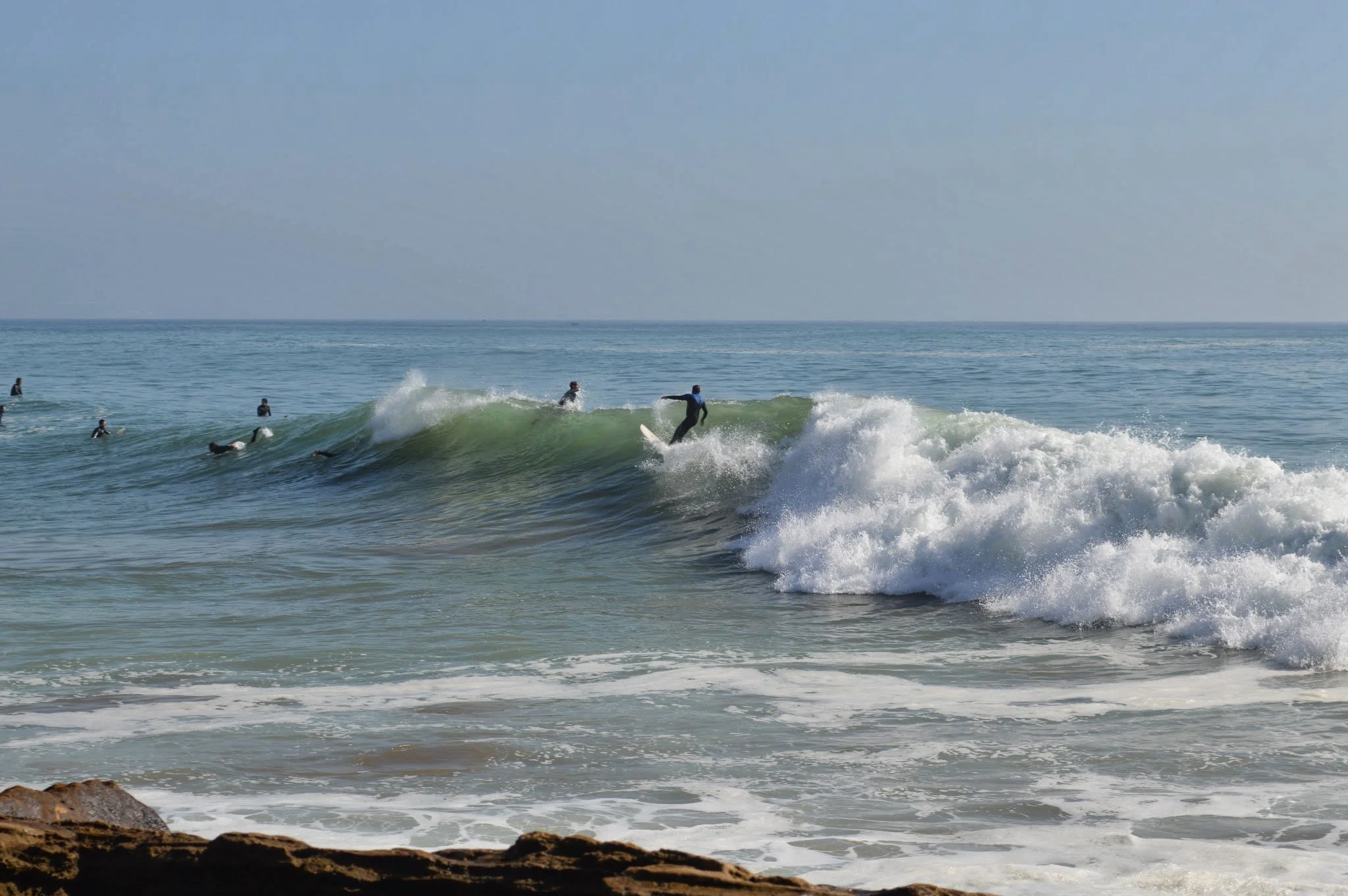 People surfing and swimming in the ocean near a rocky shore, with the sky visible in the background.