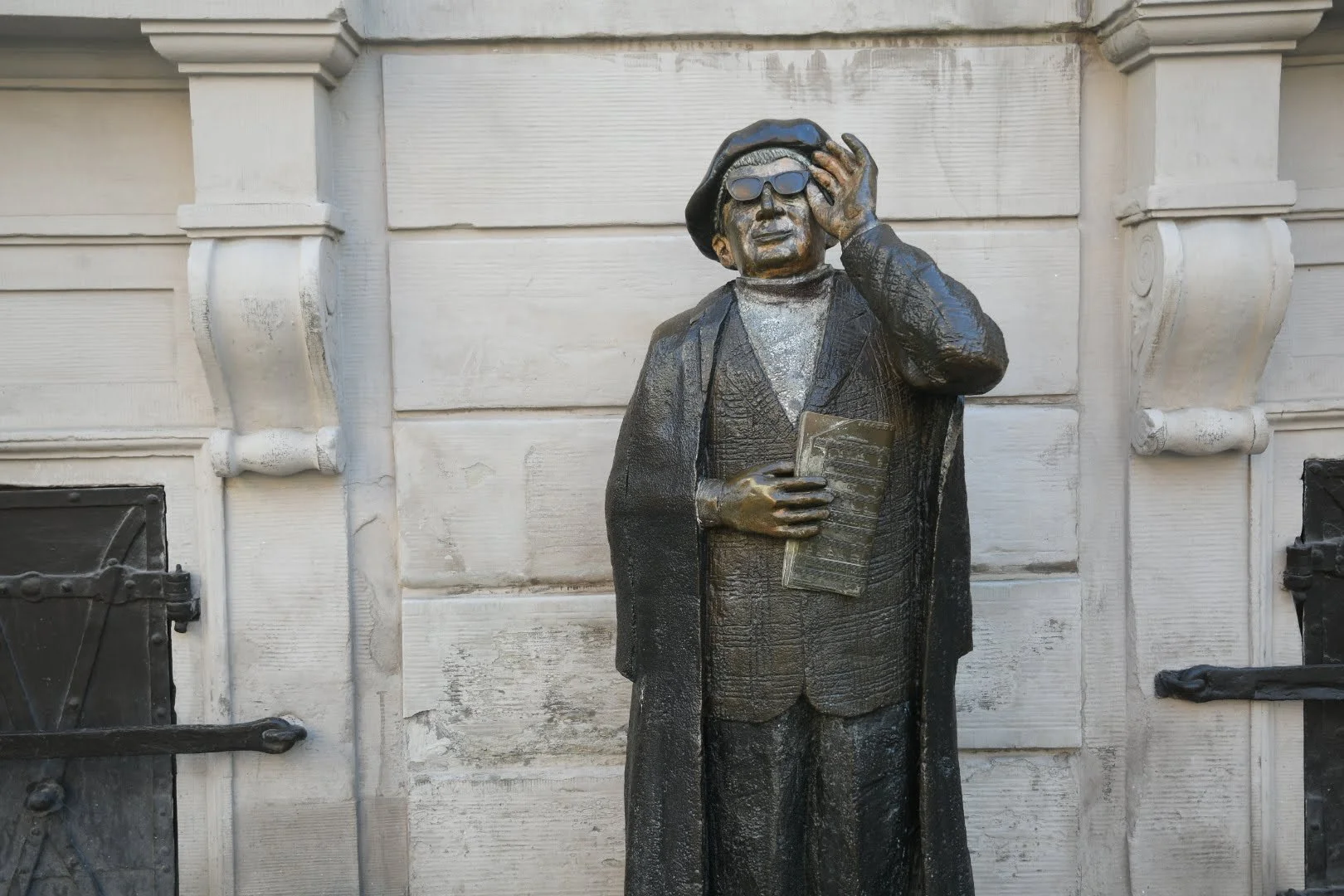 Bronze statue of a man wearing sunglasses, holding a newspaper, and adjusting his beret, standing in front of a light-colored building.