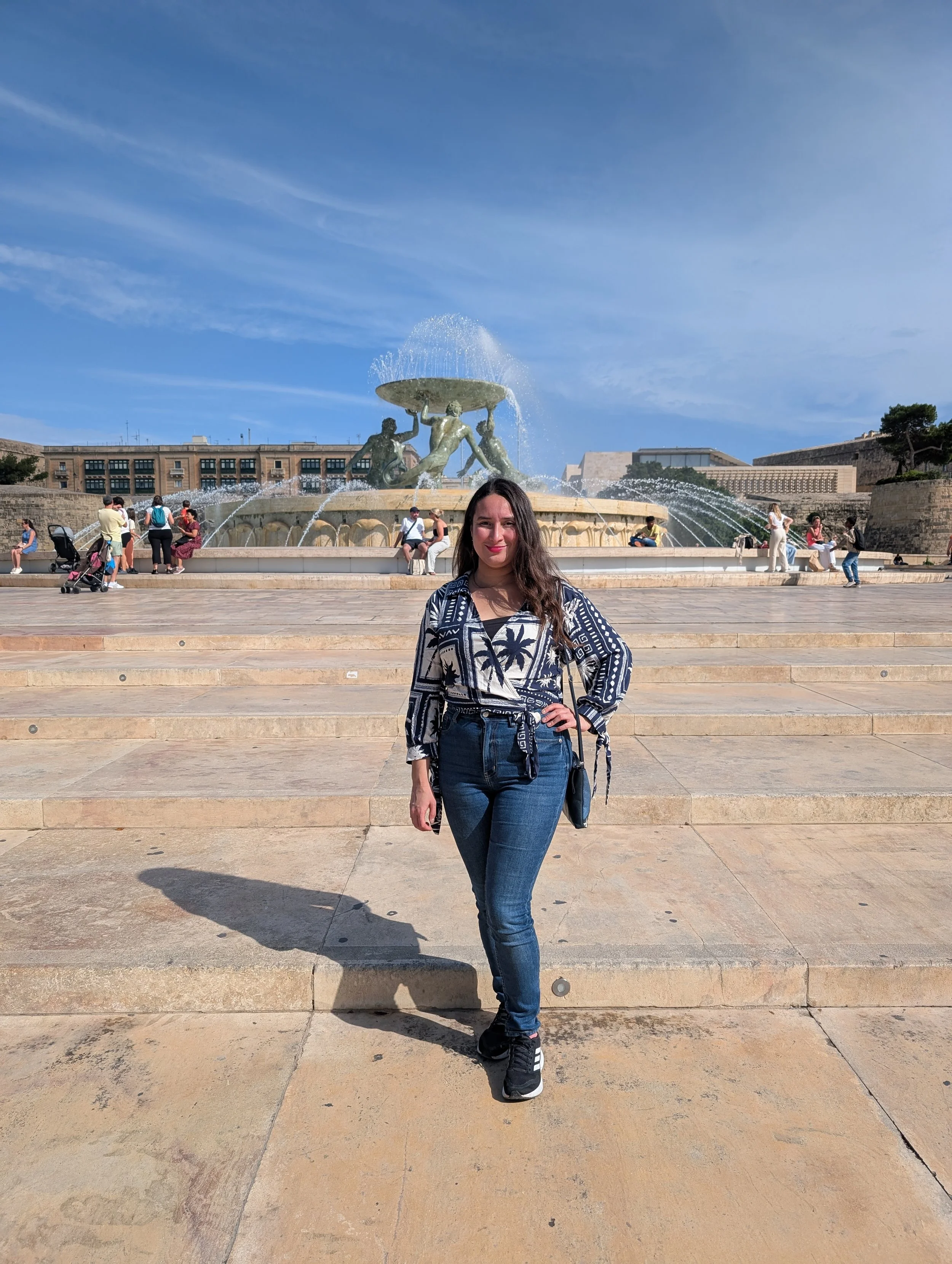 A woman with long dark hair and a patterned black and white shirt stands in front of a large fountain with statues of three figures supporting a large circular basin, with a blue sky and a few clouds in the background. Several people sit and stand ar