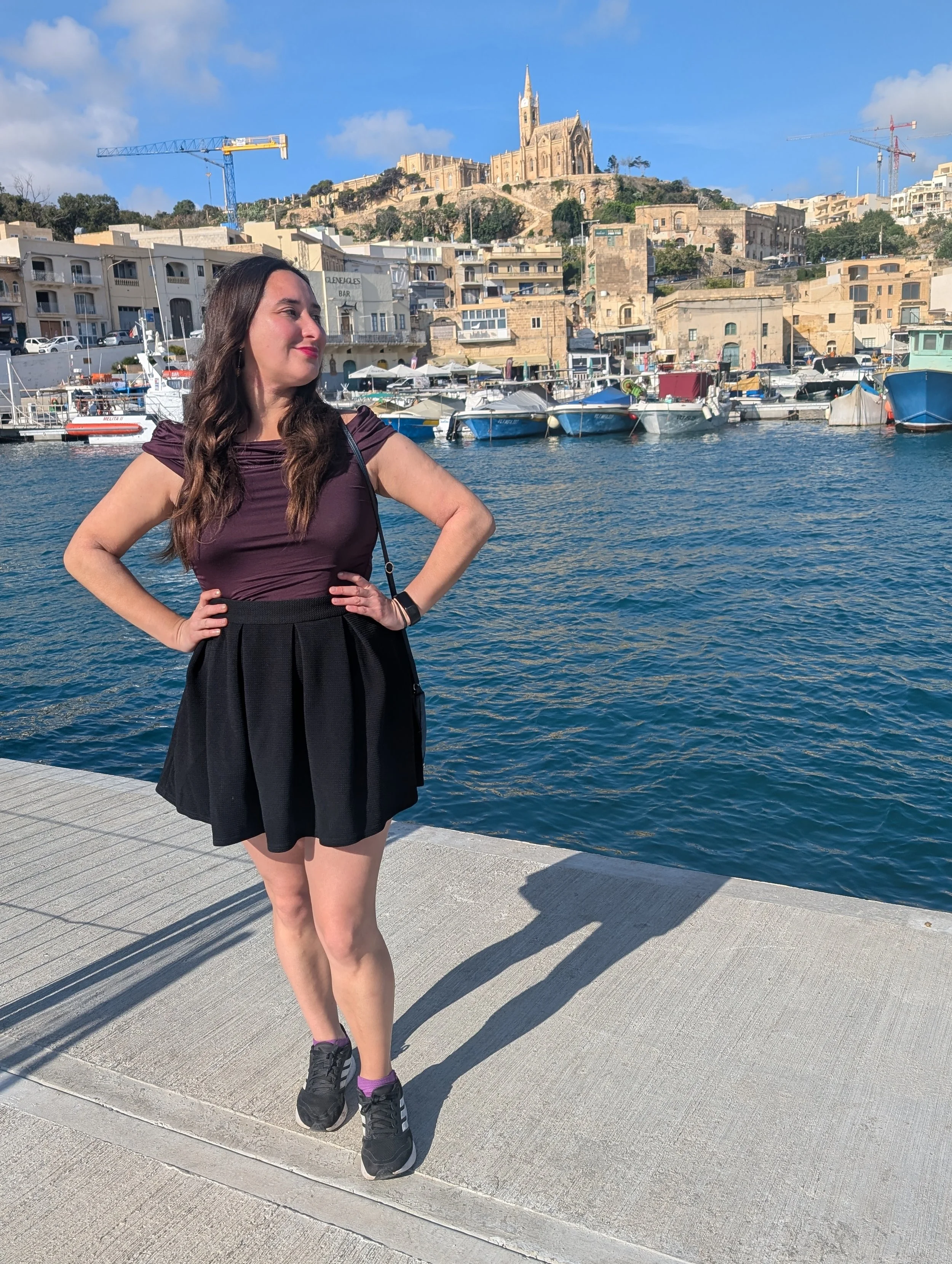 A woman with dark hair wearing a purple top and black skirt posing with hands on hips near a harbor with boats. In the background, there is a hillside with a large church and cranes under a blue sky with clouds.