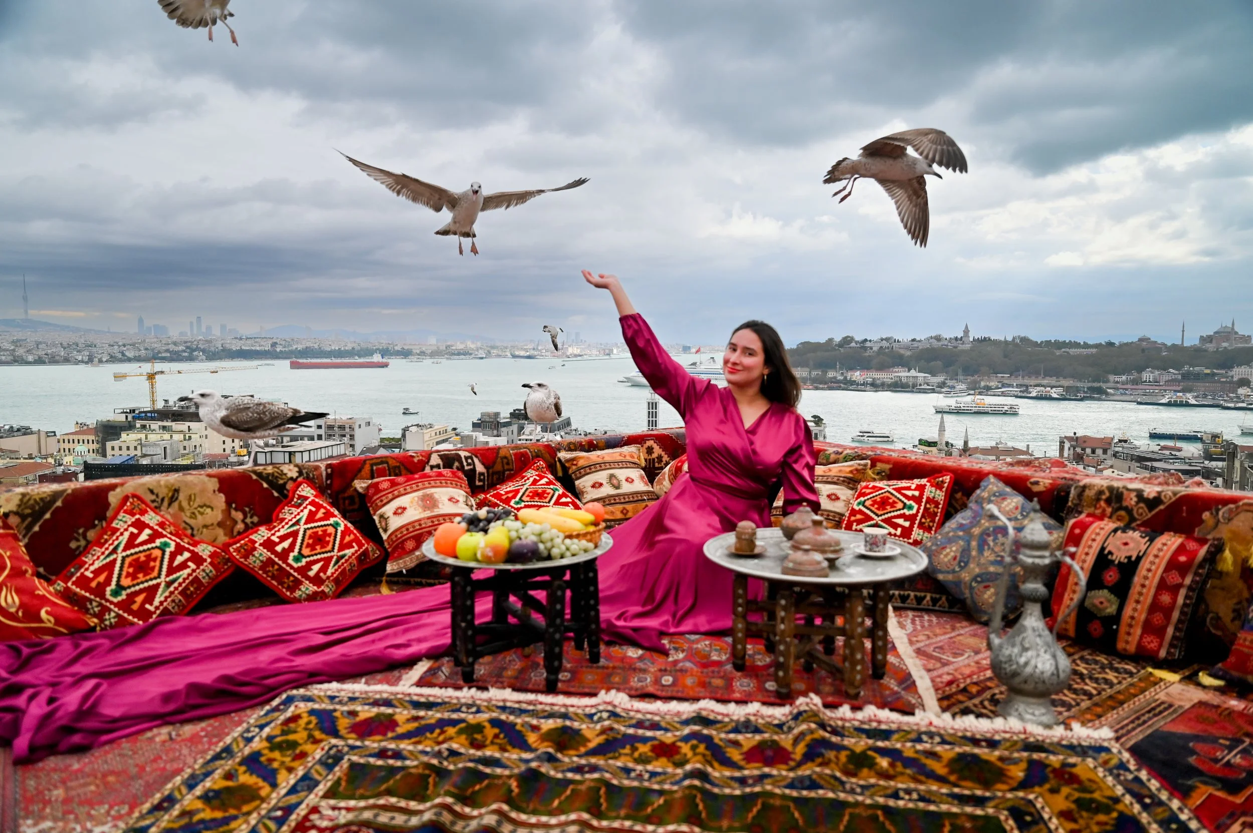 A woman in a pink dress sitting on a colorful, patterned couch with Persian-style cushions, holding her arm up while birds fly overhead near a city skyline and river, with food and drinks on a table in front of her.
