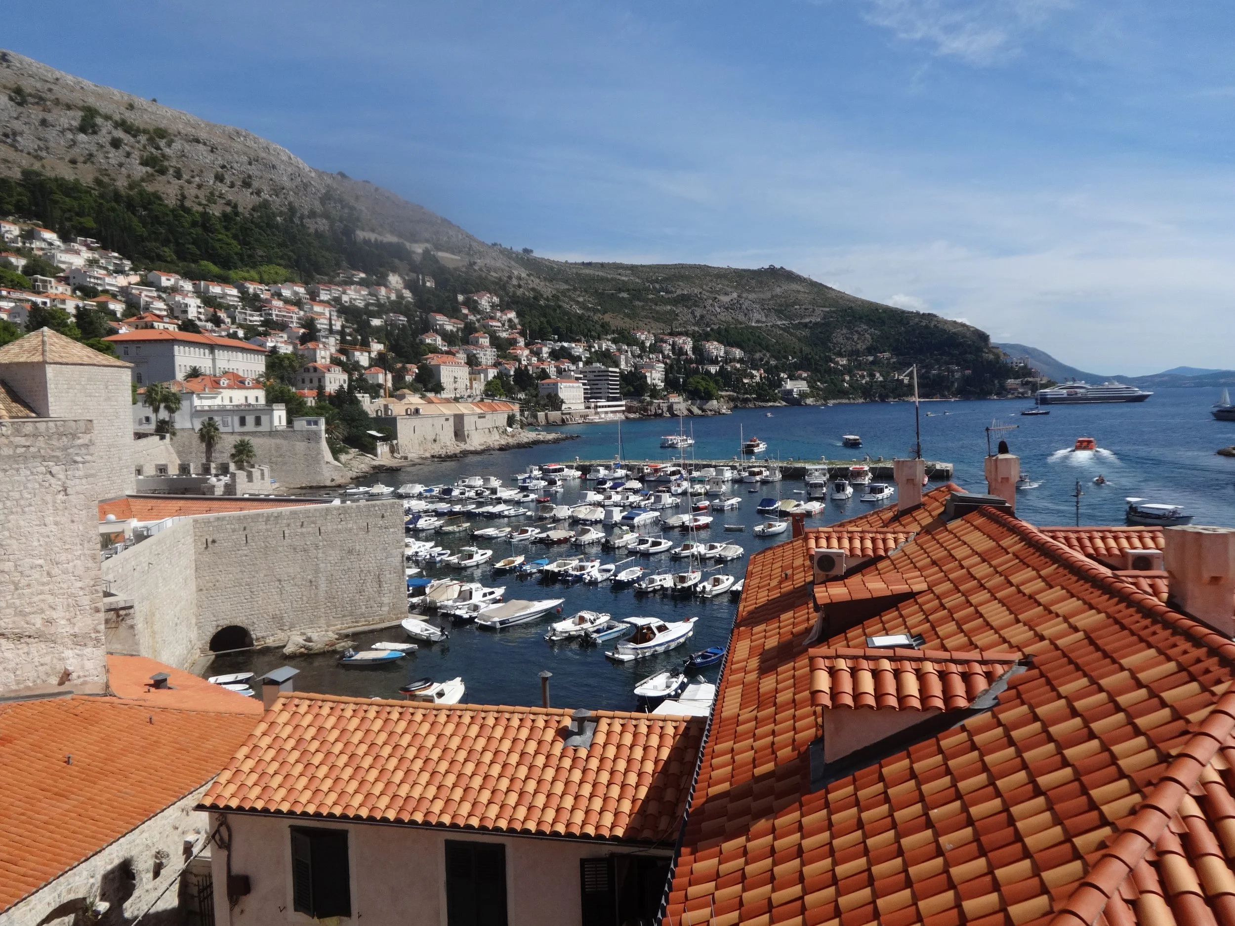 View of a coastal town with terracotta rooftops, a marina with boats, and hills in the background under a partly cloudy sky.