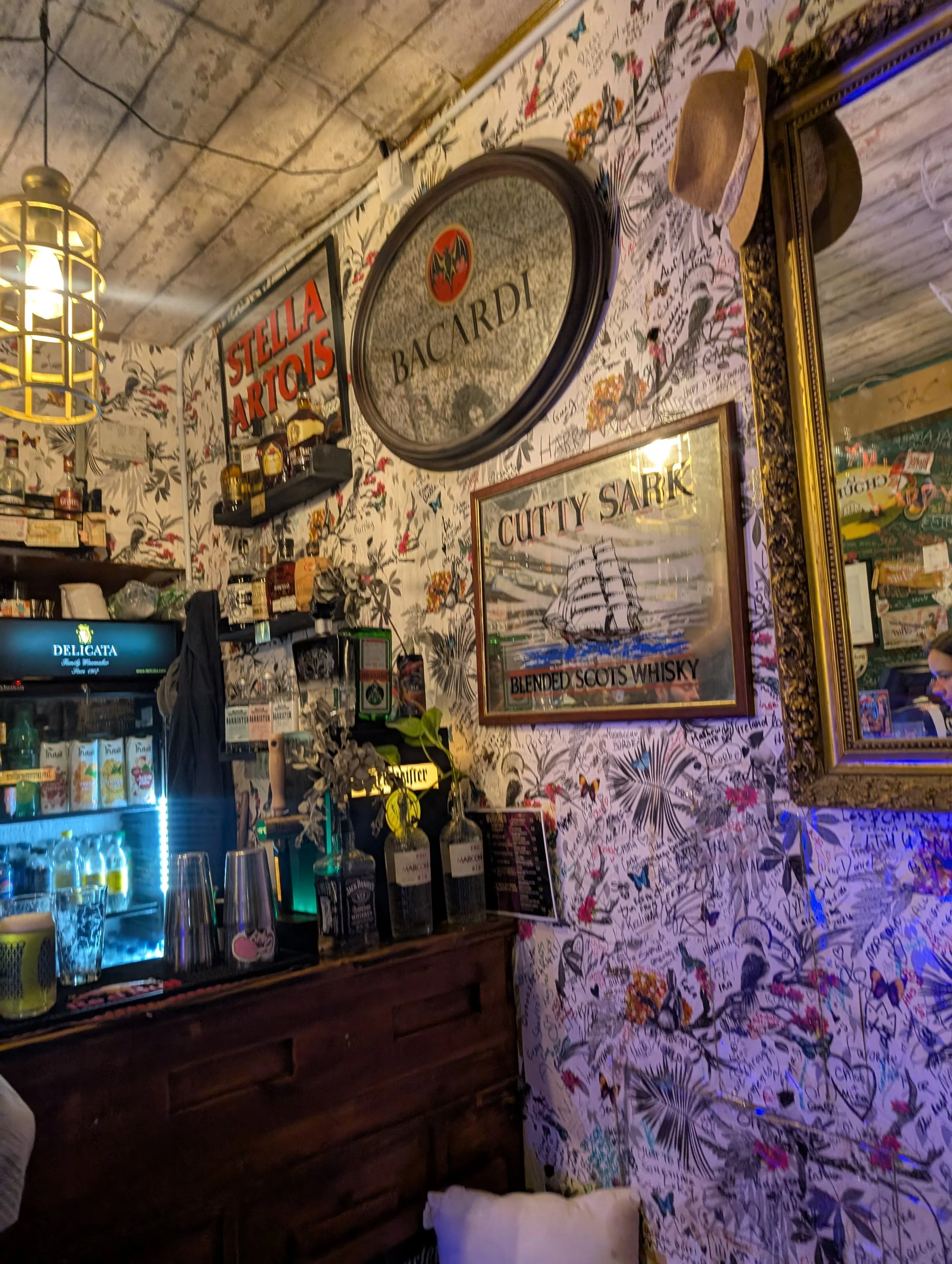 The image shows the interior of a bar or pub with a rustic, vintage style. There are various bottles of alcohol on shelves, a small TV, and decorative posters and signs, including Bacardi and Scotch whisky advertisements. The wall is decorated with f