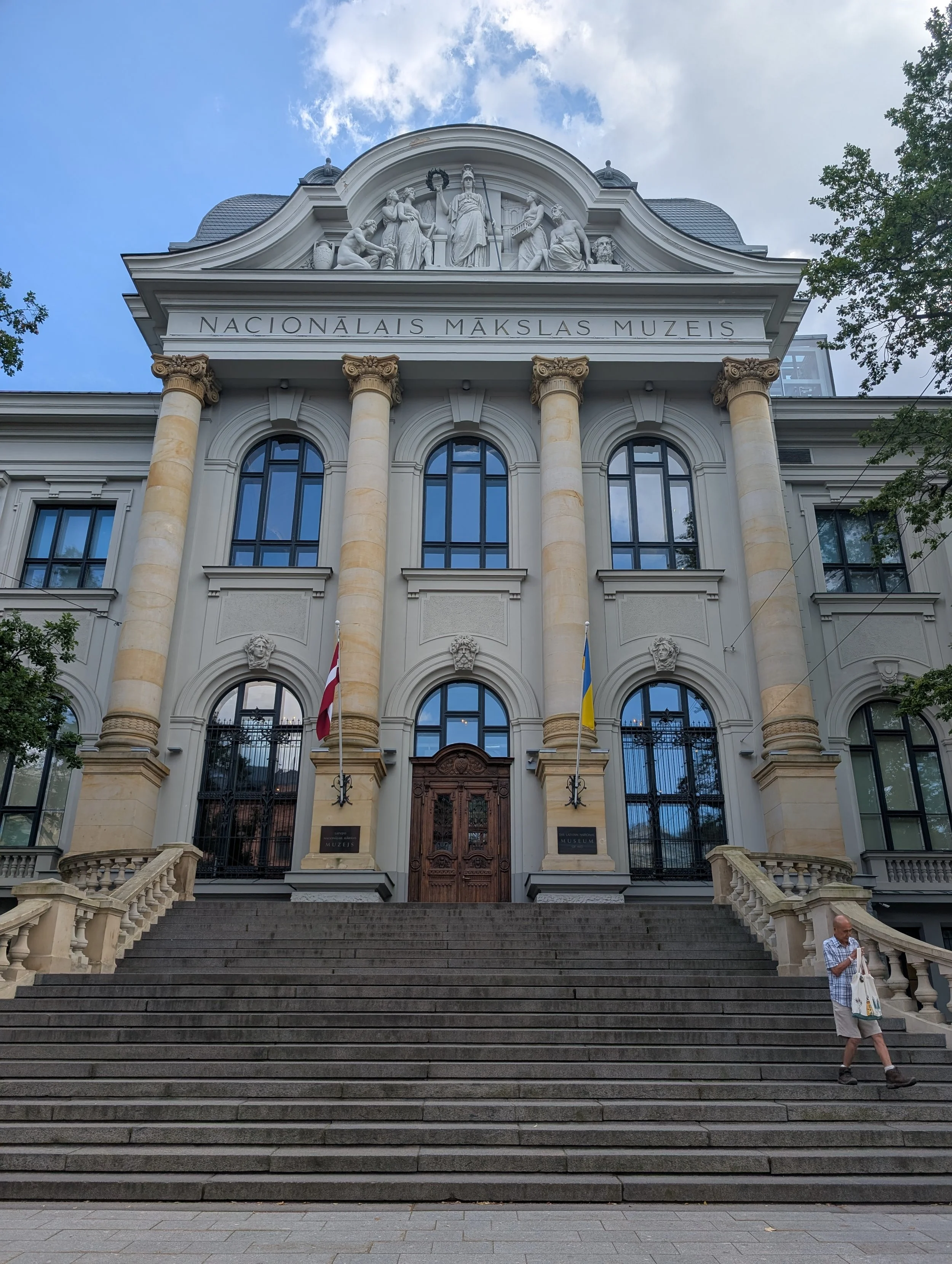 Front view of a classical style building with columns and steps, with the sign 'NACIONÁLAIS MĀKSLAS MUZEJS' on the facade, and a man walking down the stairs on the right.