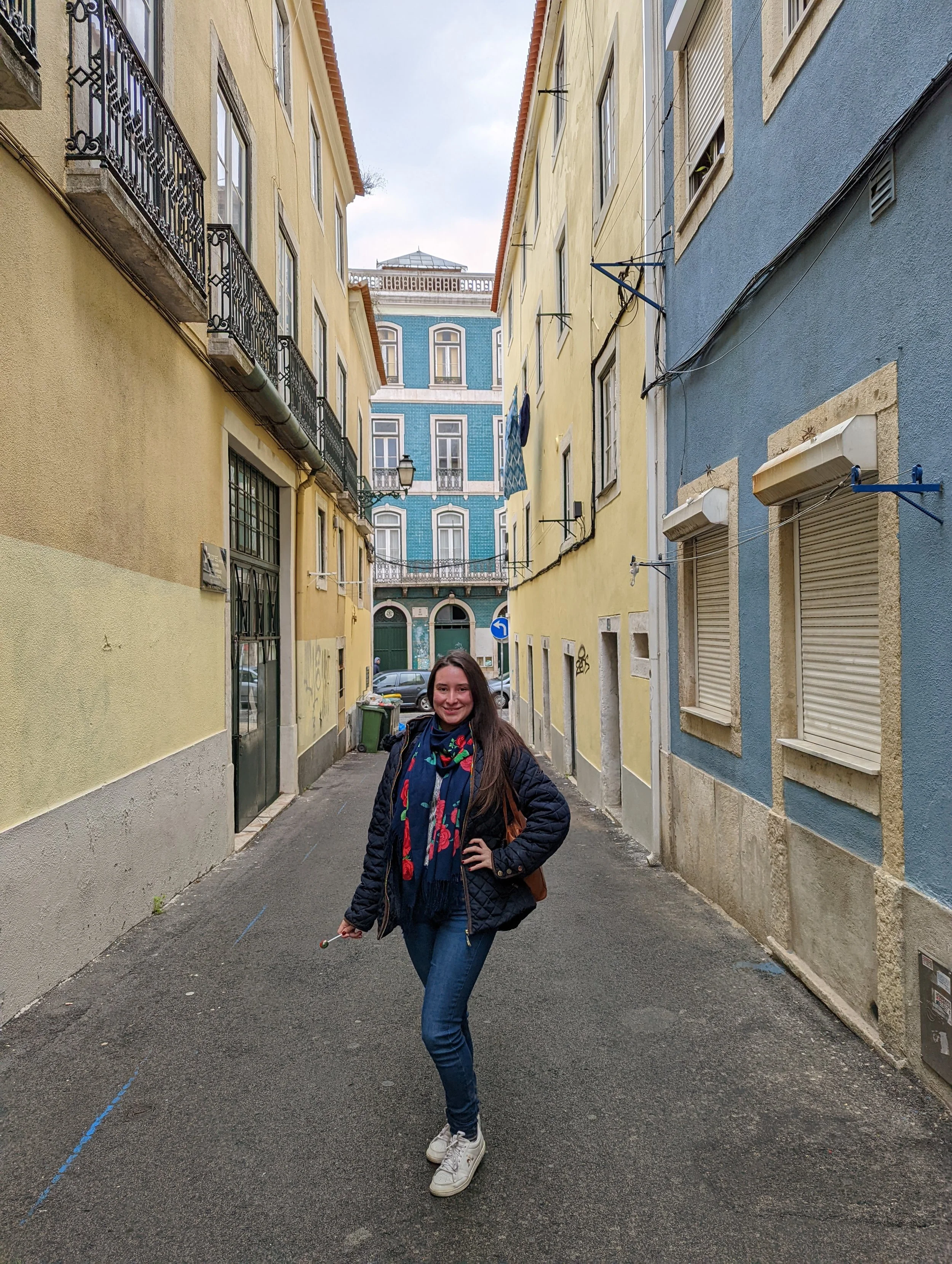 A smiling woman wearing a navy jacket, blue jeans, white sneakers, and a colorful scarf, standing in a narrow European street with pastel-colored buildings on either side and a man walking in the background.