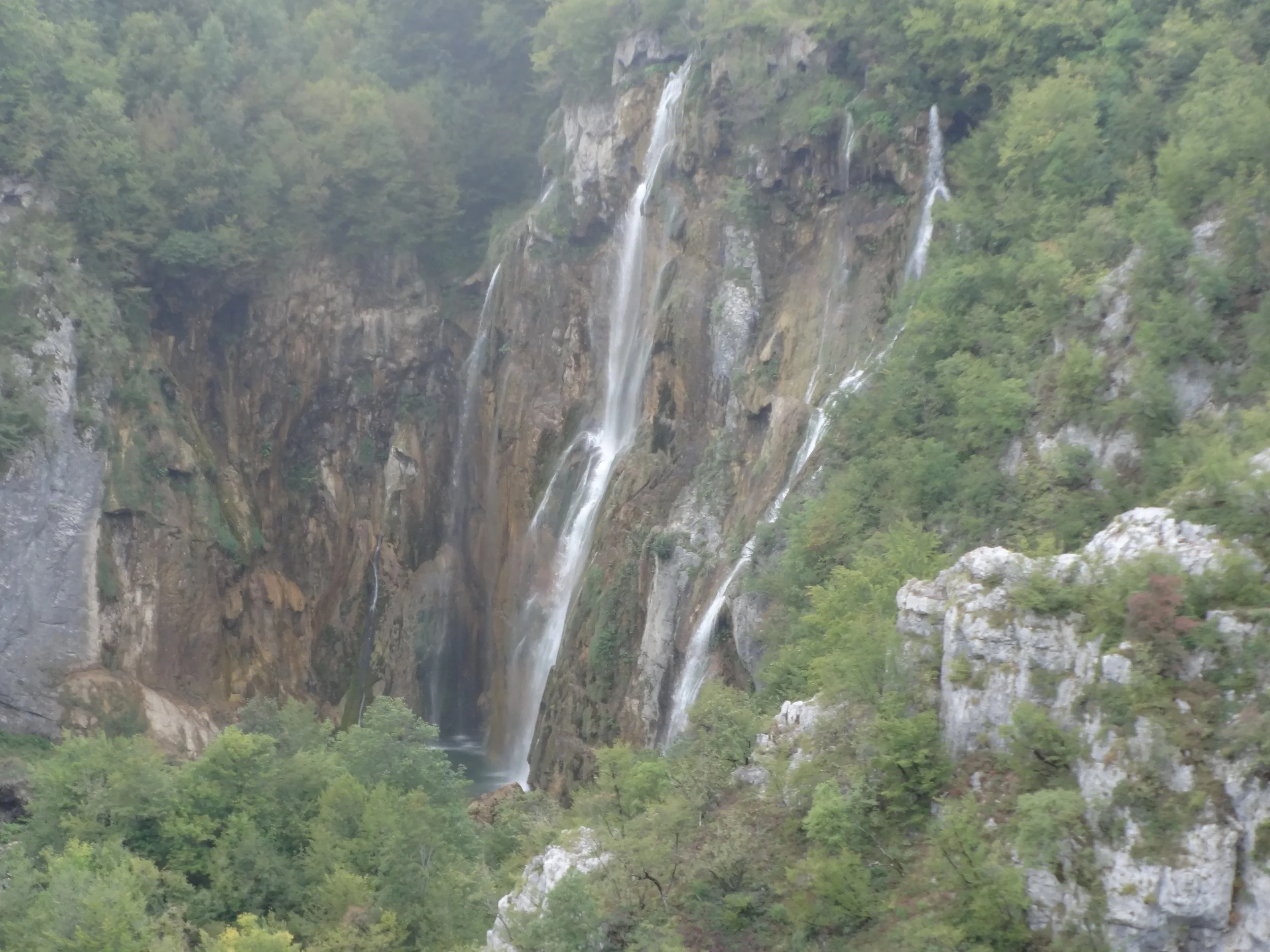 Waterfalls cascading down a rocky cliff surrounded by lush green trees and vegetation.