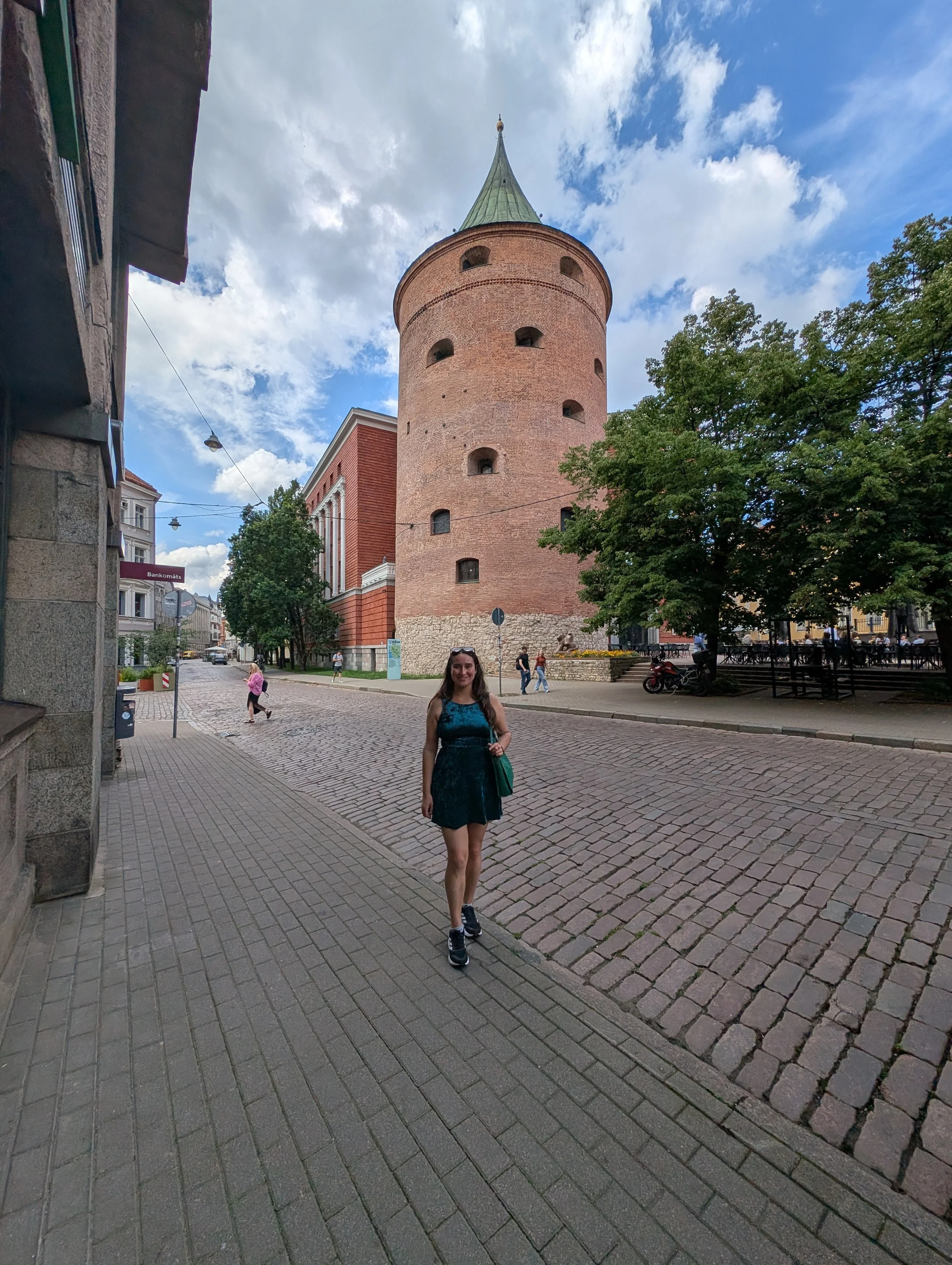 A young woman wearing a blue dress and sneakers walking along a cobblestone street in front of a historic brick tower with a pointed green roof. Several people are visible in the background, along with trees and buildings.