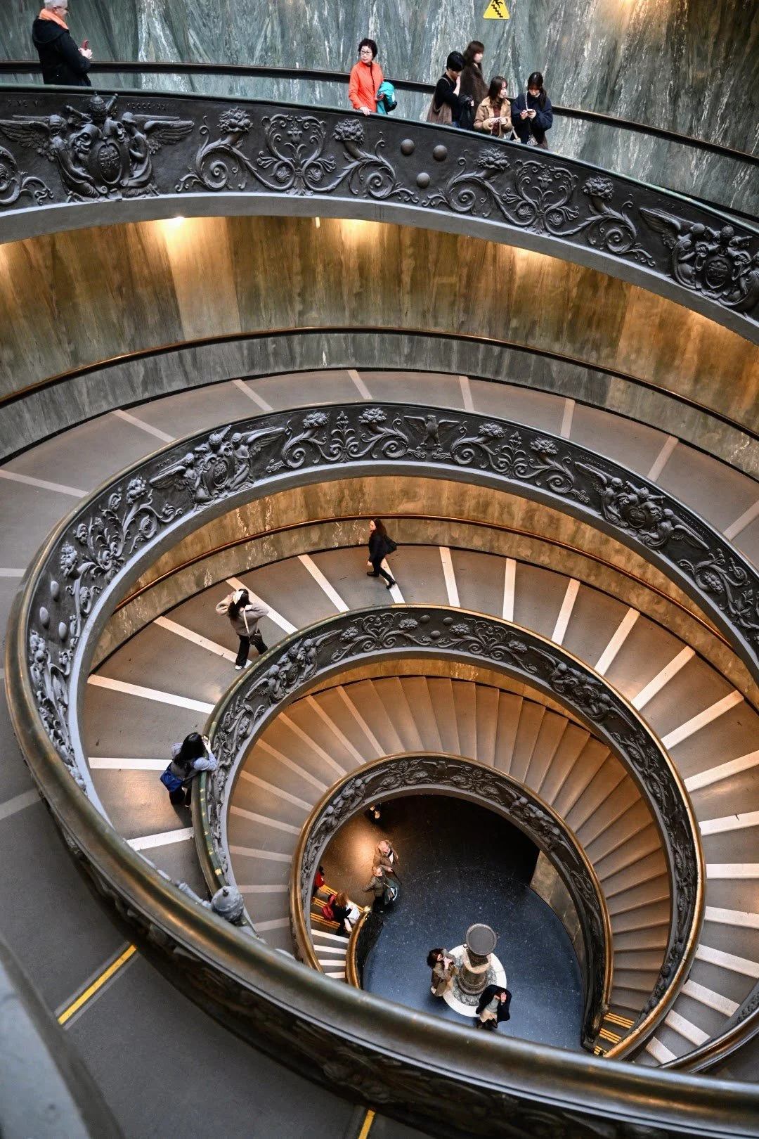 Spiral staircase viewed from above, with people walking on the stairs and decorative black railing with ornate floral patterns.