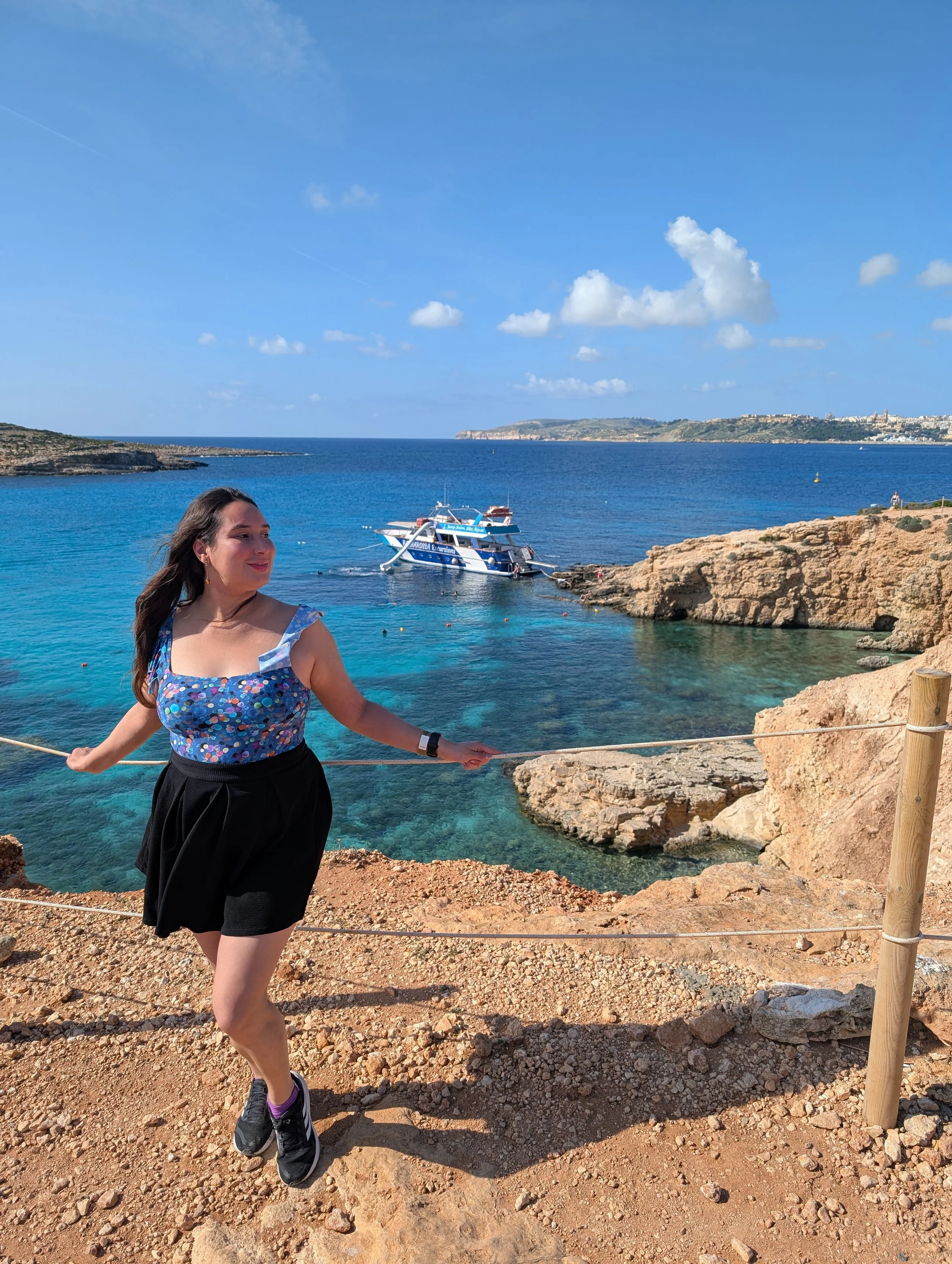 A woman standing on a rocky seaside with clear blue water and a boat in the background, under a partly cloudy sky.