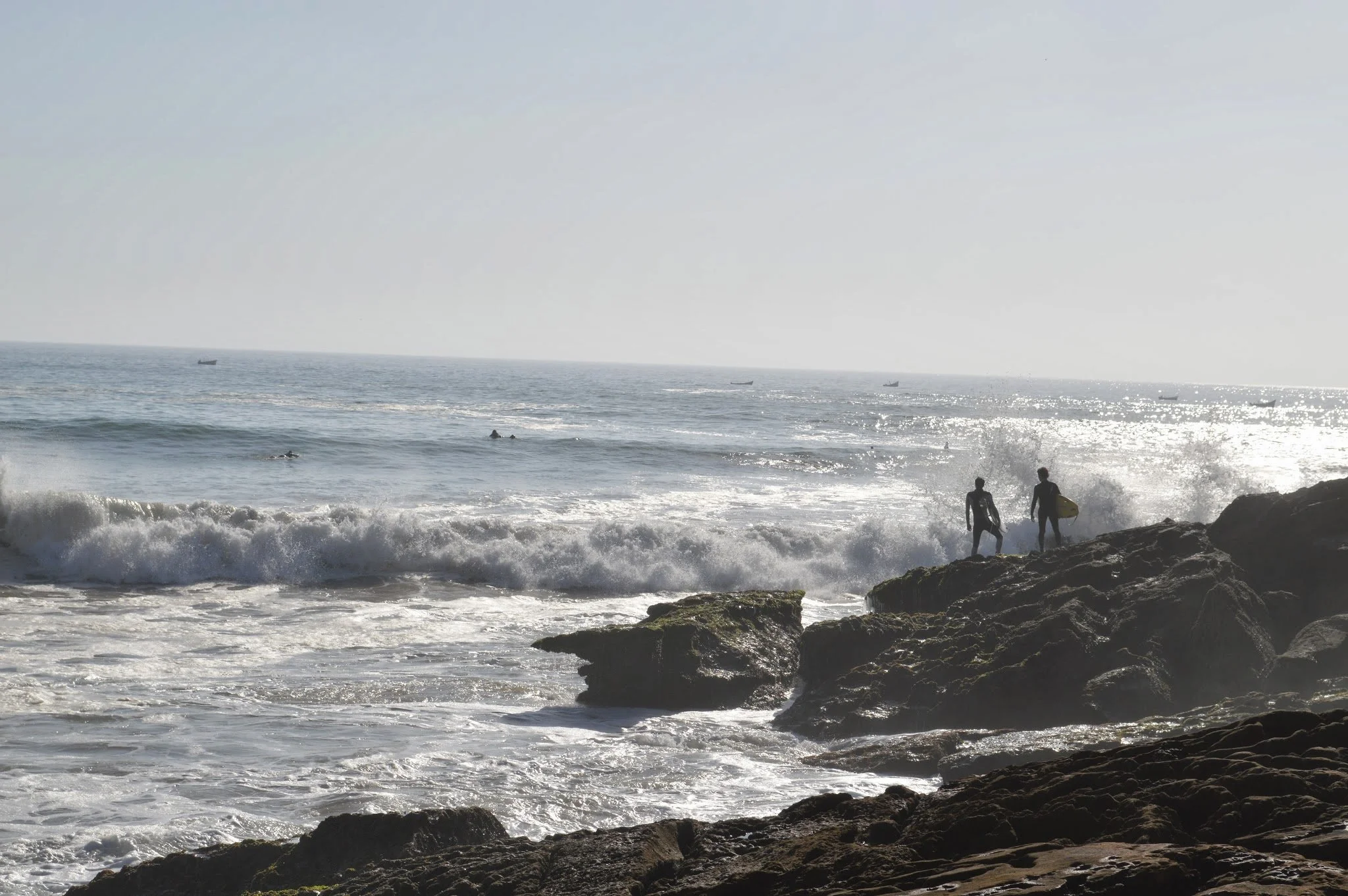 Two surfers carrying surfboards walk along rocky shore while waves crash at their feet, with boats sailing on the horizon under a bright, hazy sky.
