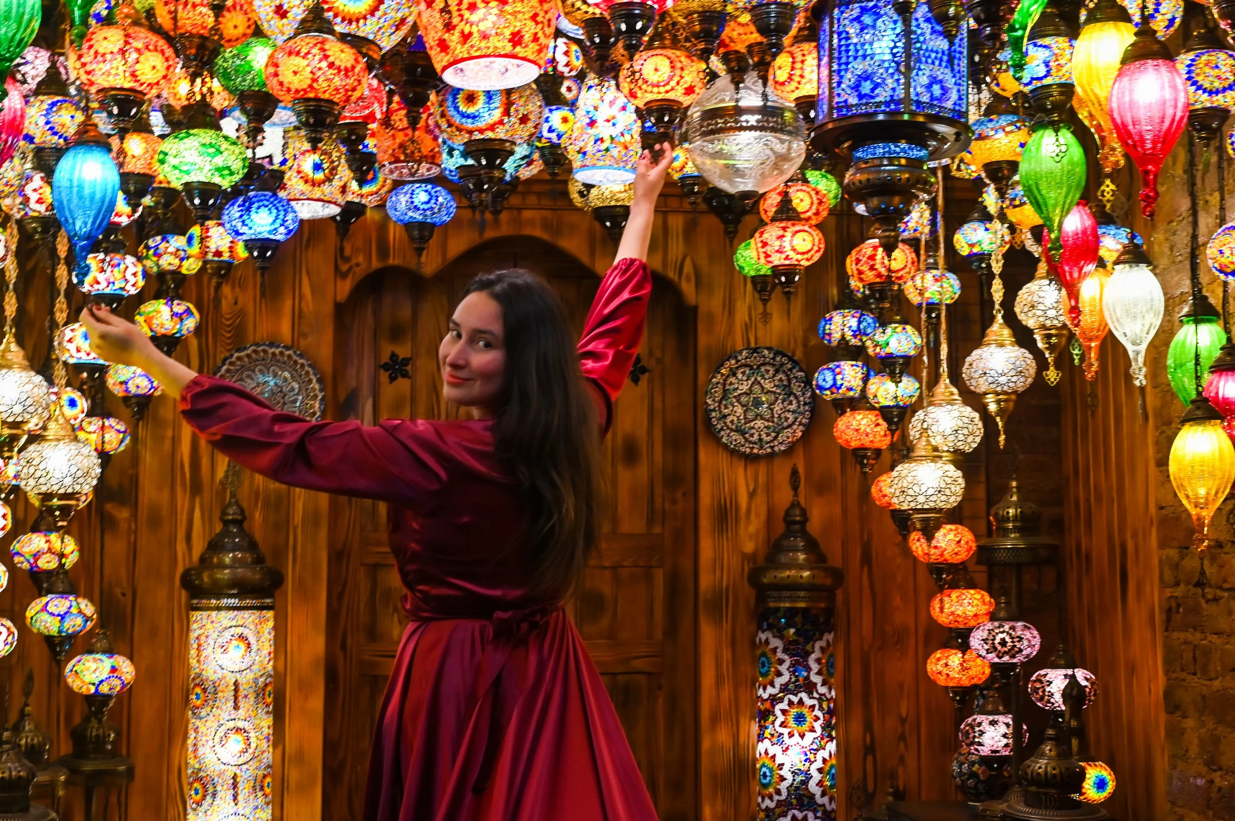 A woman with long dark hair wearing a maroon dress surrounded by colorful hanging mosaic lamps in various shapes and sizes, inside a store or marketplace with wooden walls.