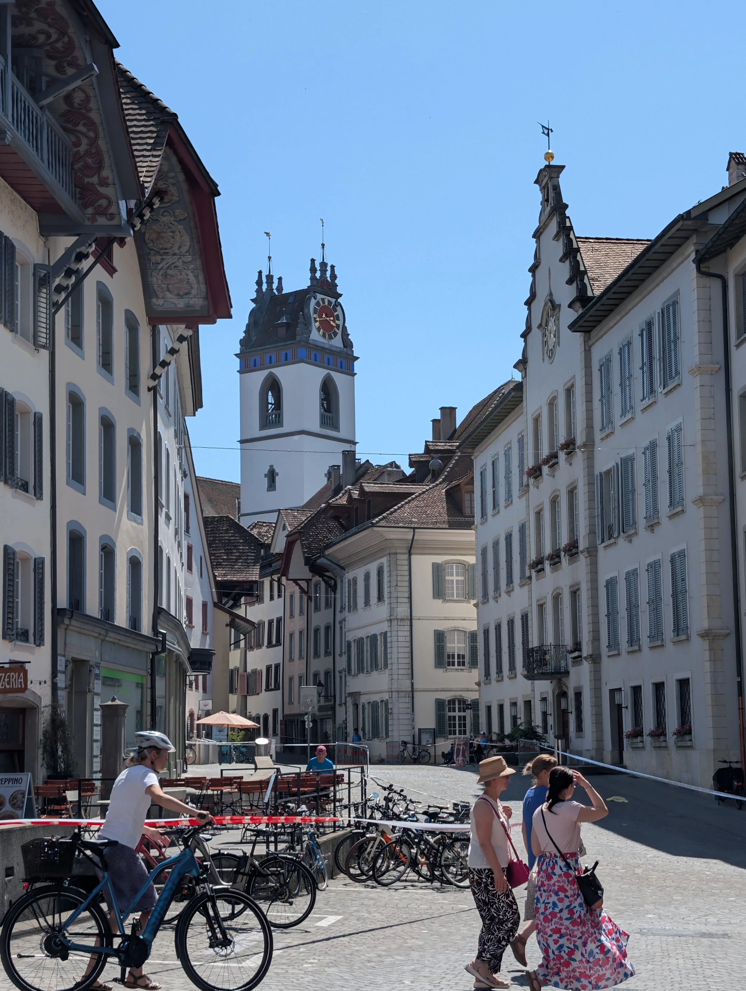 People walking and biking on a pedestrian street in a European city with historical buildings and a clock tower.