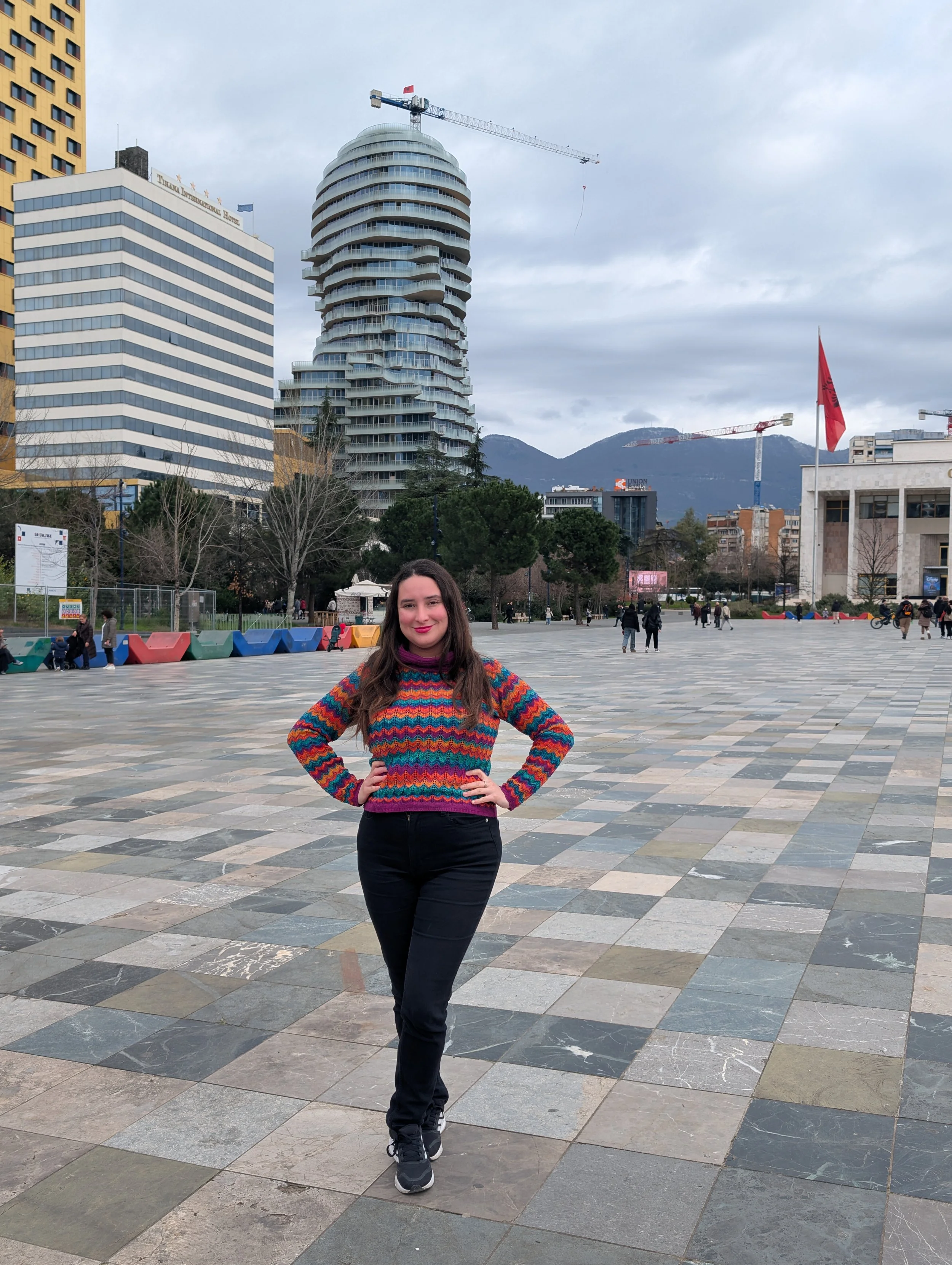 Tamara with long dark hair, wearing a colourful striped sweater and black pants, standing with hands on hips in an urban plaza with high-rise buildings, trees, and a mountain range in the background.