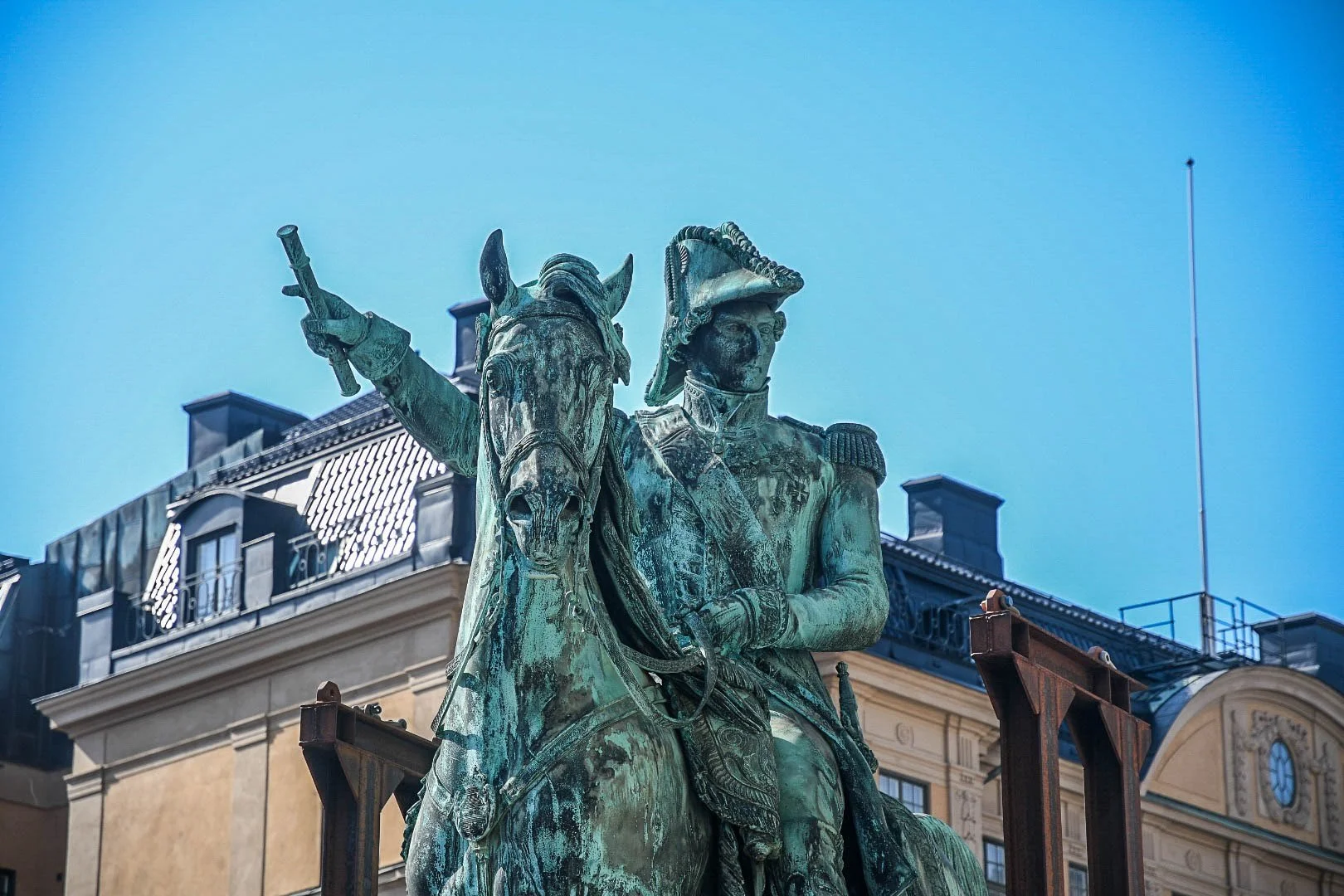 Bronze statue of a military leader on horseback, holding a baton, with buildings and a clear blue sky in the background.