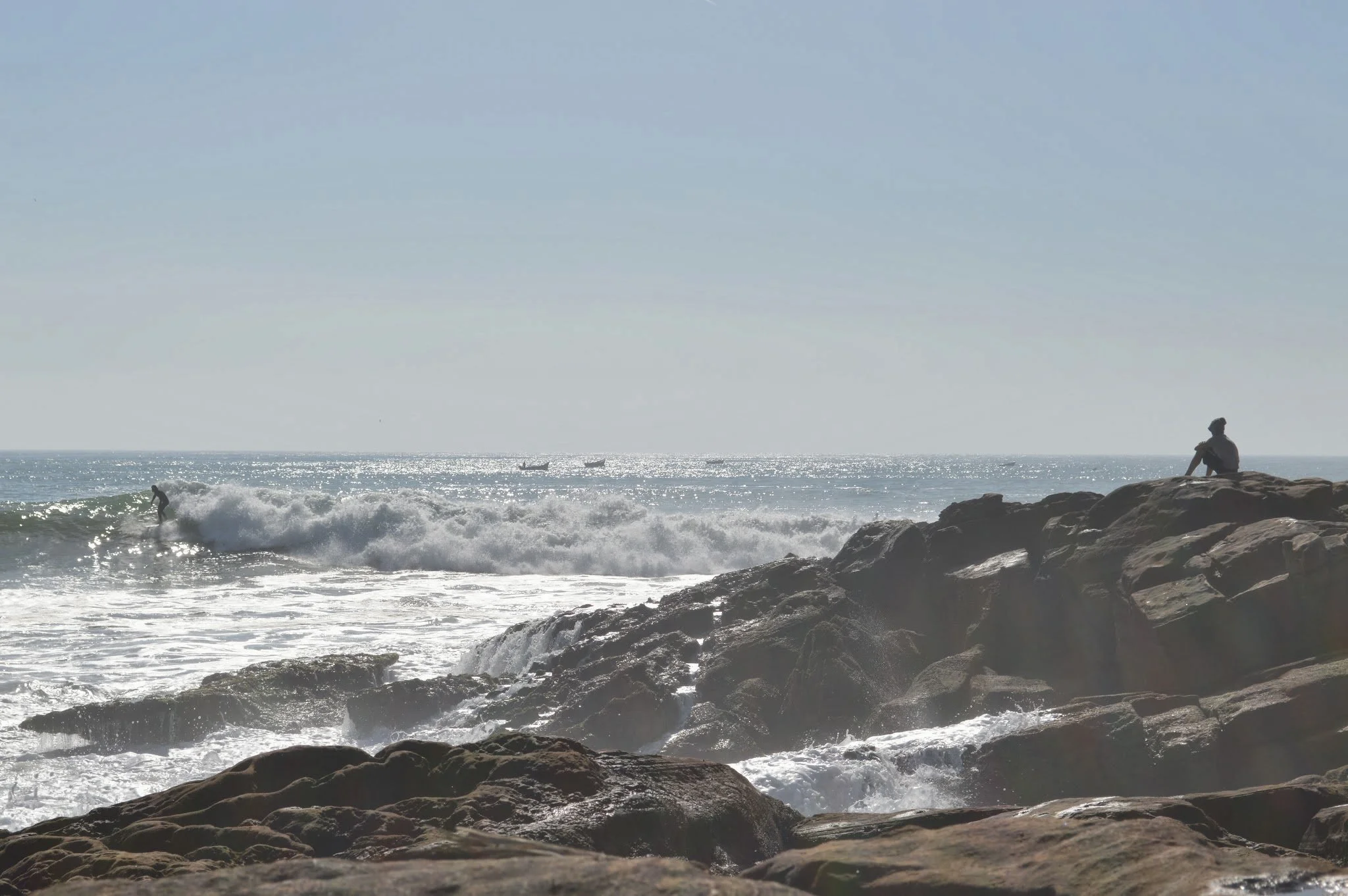 A person is sitting on rocks by the ocean, watching waves and boats on the water, with a clear sky above.