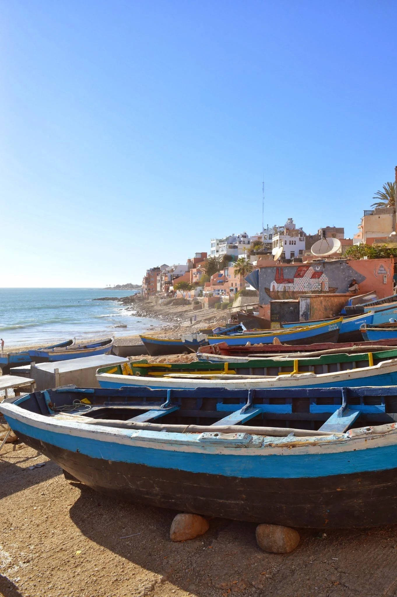 A coastal scene with numerous colourful fishing boats resting on the sandy shore and a hillside populated with white and pastel-colored buildings, some with satellite dishes and antennas, overlooking the ocean under a clear blue sky.