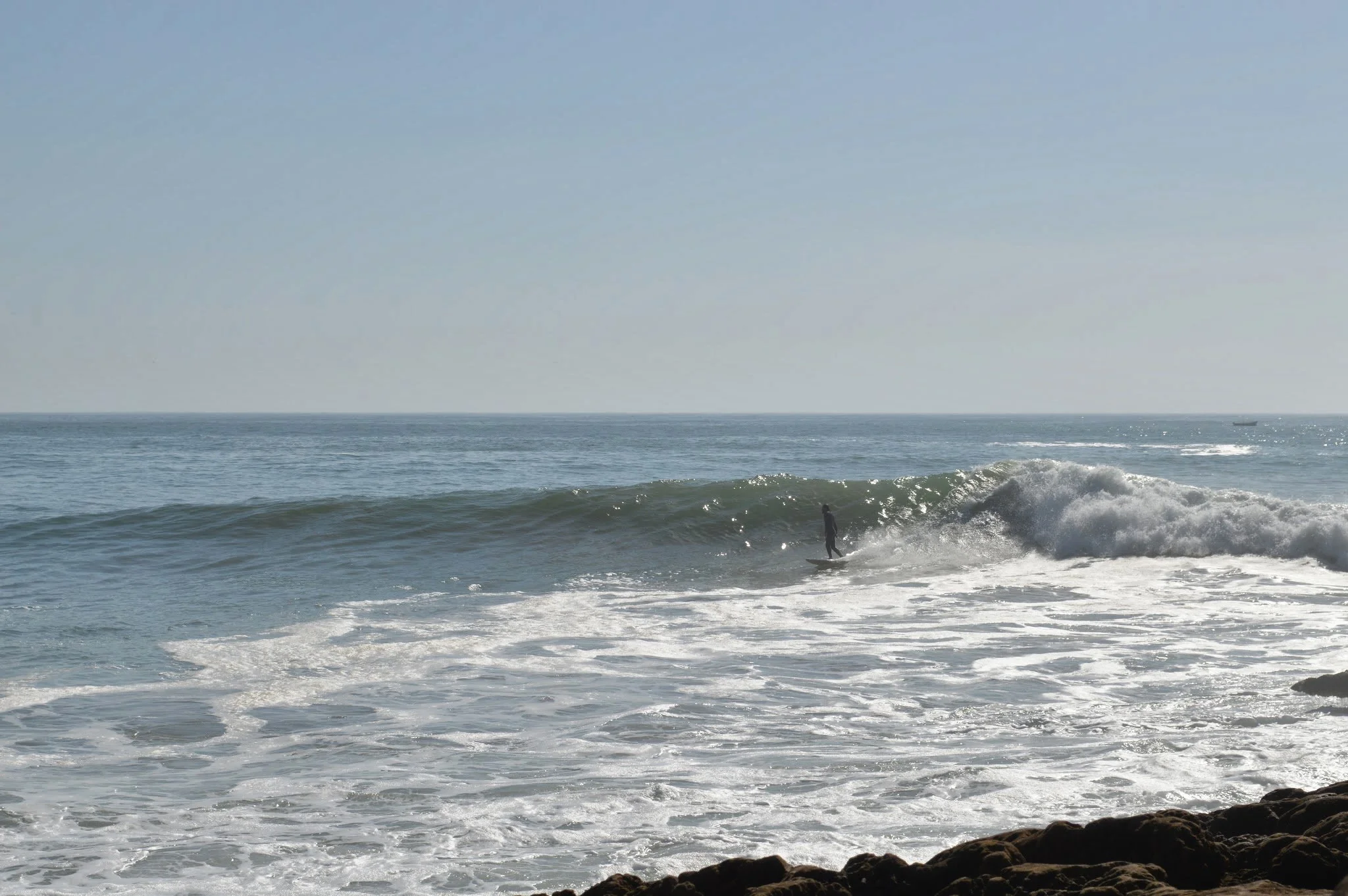 A person surfing on a wave in the ocean near rocks with a boat in the distance on a clear day.