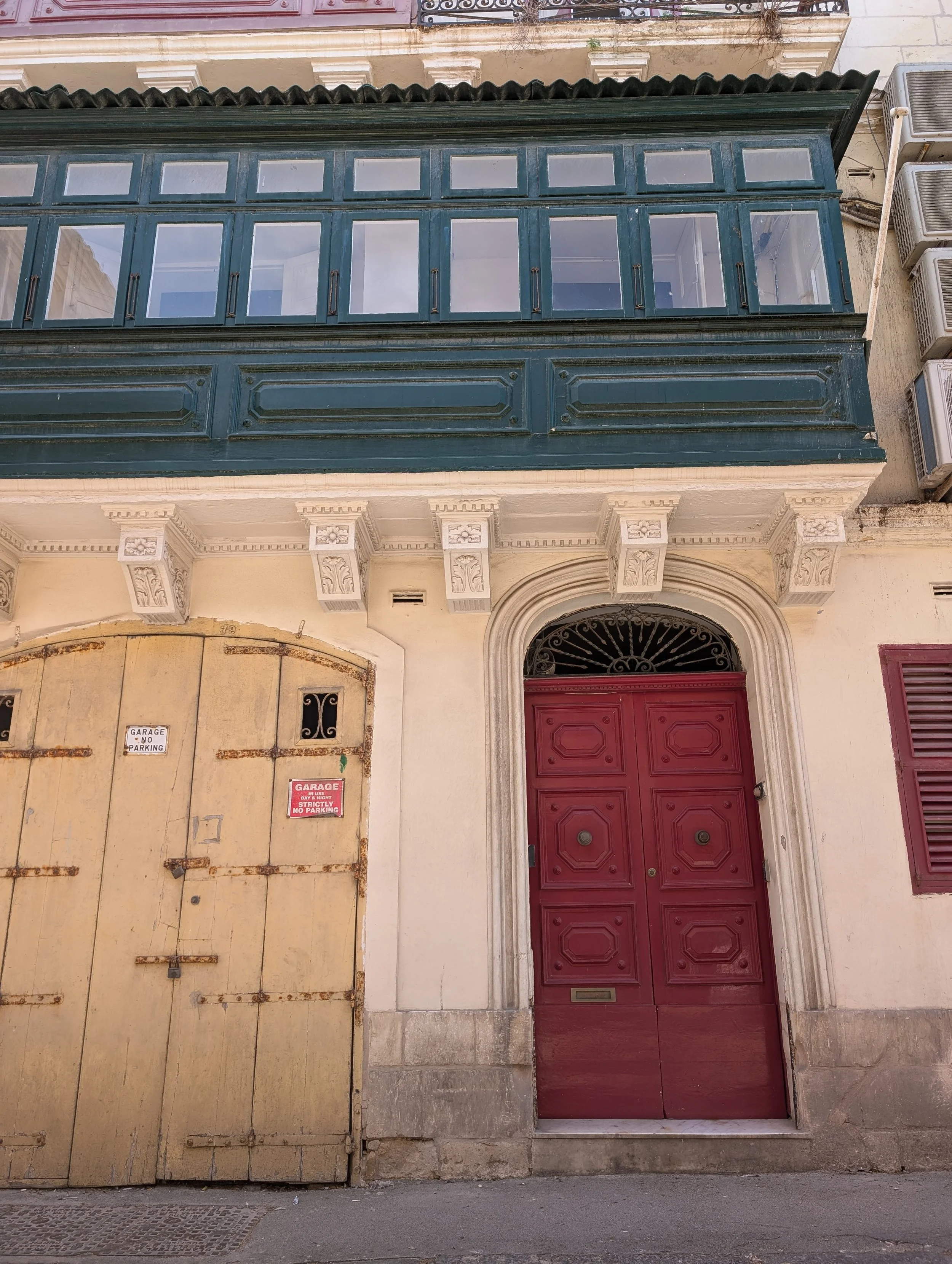 A building facade features a large, ornate wooden door painted red with rotary door handles, framed by decorative stonework, including floral motifs. To the left, there is a large, old yellow metal garage door with multiple signs indicating no parkin
