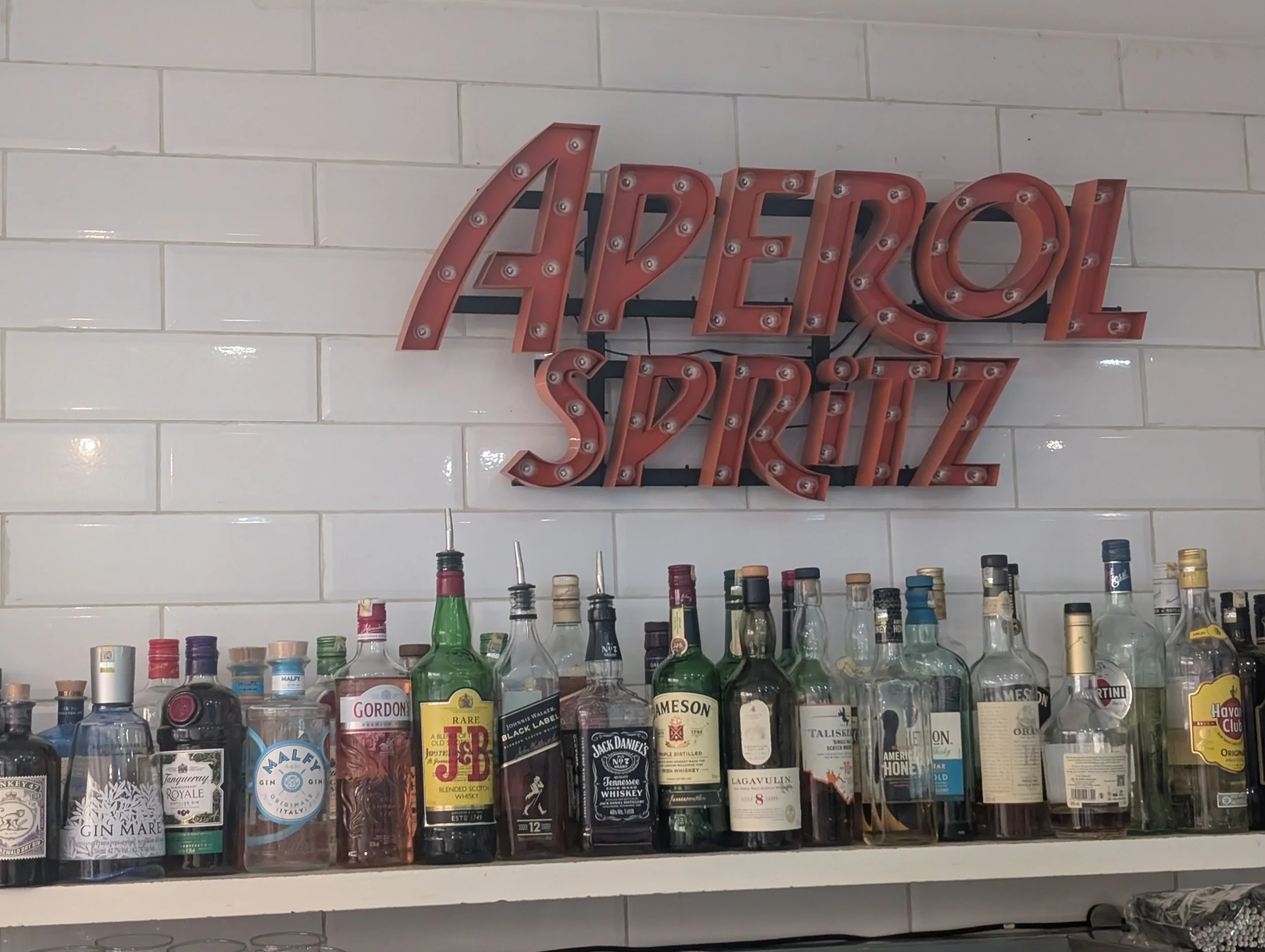 A wall-mounted sign that reads 'APEROL SPRITZ' in illuminated vintage style lettering with light bulbs outlining the letters, above a shelf with various bottles of alcohol.