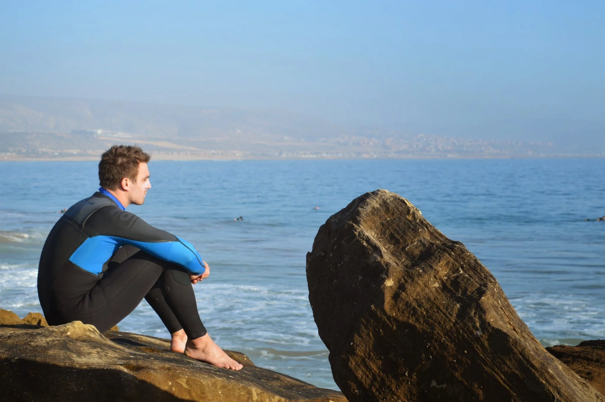 A young man dressed in a wetsuit sitting on rocks by the ocean, looking contemplative with a large rock in front of him, with distant land and mountains in the background.
